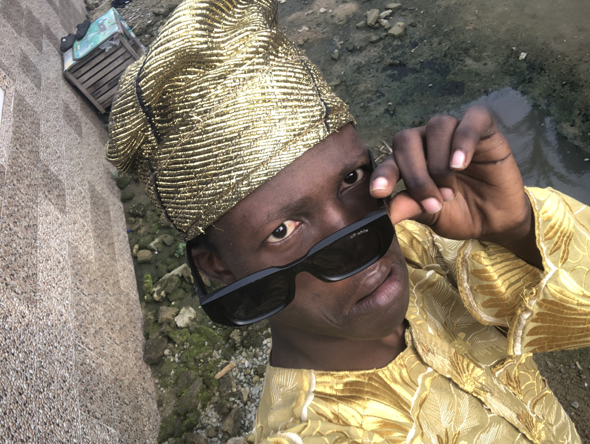 A close-up, high-angle shot shows a person wearing a gold headwrap and ornate gold clothing, positioned in the city of Ado, Nigeria. The subject is looking upwards, and their right hand is adjusting dark sunglasses. The background consists of a rough, textured wall with small stones and some moss, leading down to an area with loose soil, rocks, and what appears to be murky water. A portion of a wooden crate with a green surface is visible in the upper right corner of the image. The lighting suggests it is daytime.