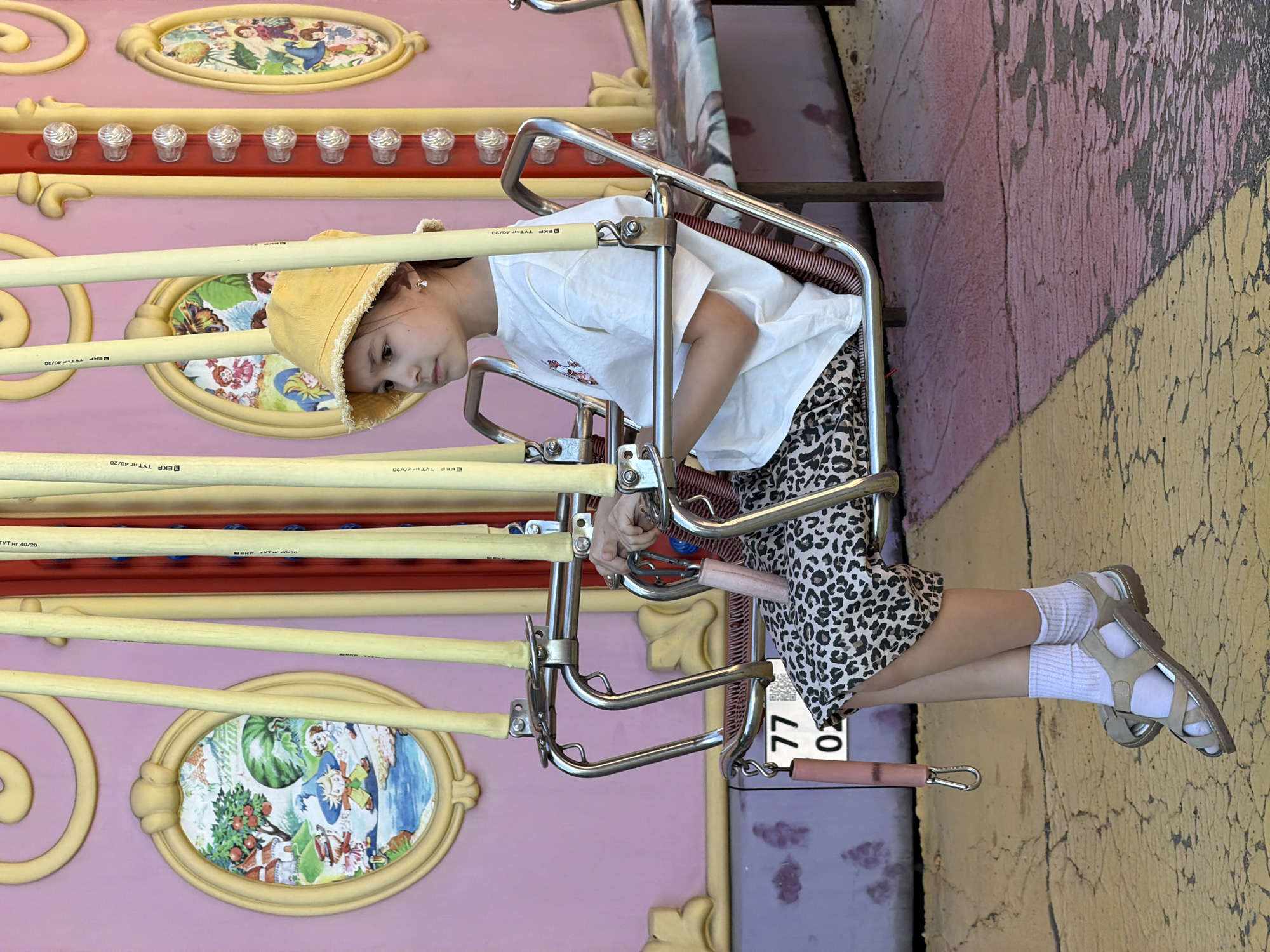 This image depicts a young girl seated on a stationary swing ride within an amusement park setting in Moscow, Russia. The girl, appearing pre-adolescent, has light-colored skin and dark hair, and wears a yellow bucket hat, a loose-fitting white short-sleeved t-shirt, and leopard print shorts. She is wearing white ankle socks and light brown, multi-strap sandals. Small stud earrings are visible. Her hands rest on the chrome safety bar of the swing, with her right hand holding a pink cylindrical object. She gazes towards the left of the frame with a neutral expression. The swing seat features a red ribbed backrest, and the vertical support poles are yellow and bear the text "EKF TYT-H 40/20". In the background is a large, pink-colored wall, part of the ride structure, adorned with yellow decorative scrolls and several yellow-framed oval panels containing whimsical painted scenes featuring cartoon characters, landscapes with houses, trees, and water. A vertical red panel with multiple clear, bulb-like lights or decorative studs is also visible on the pink wall. A white tag displaying "77 02" and a QR code is partially visible, hanging from the left side of the swing mechanism. Another pink cylindrical object with a metal clip hangs from the same side. The ground is paved and shows an area of yellow transitioning to faded pink/purple, with visible cracks and wear. The scene is illuminated by natural daylight.