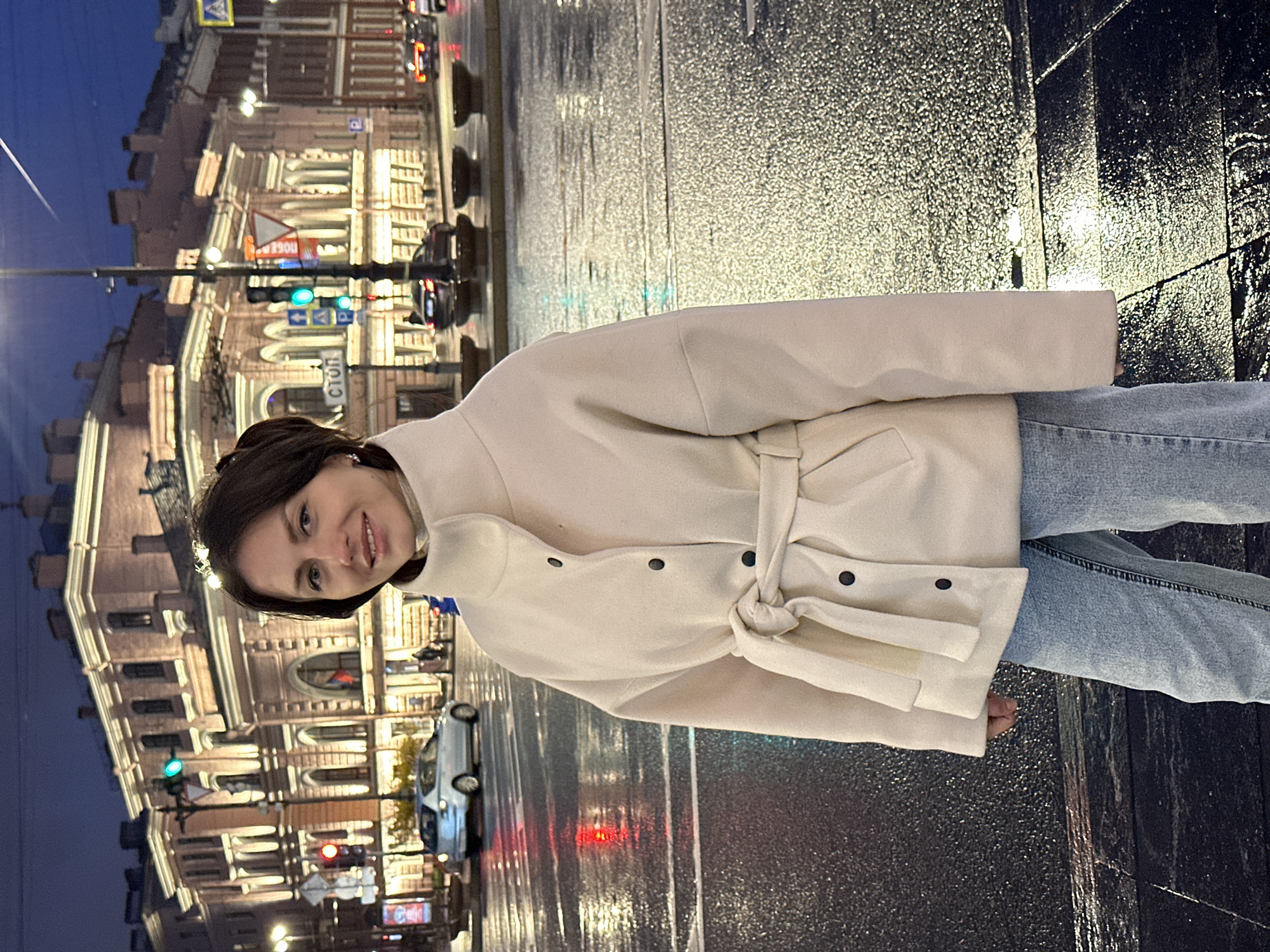 A woman stands on a wet street at night in Saint Petersburg, Russia. She is wearing a cream-colored coat with a belt and light blue jeans. The street is dark and glistening with reflections. Behind her, there is a building with illuminated windows and traffic lights. The sky is dark.