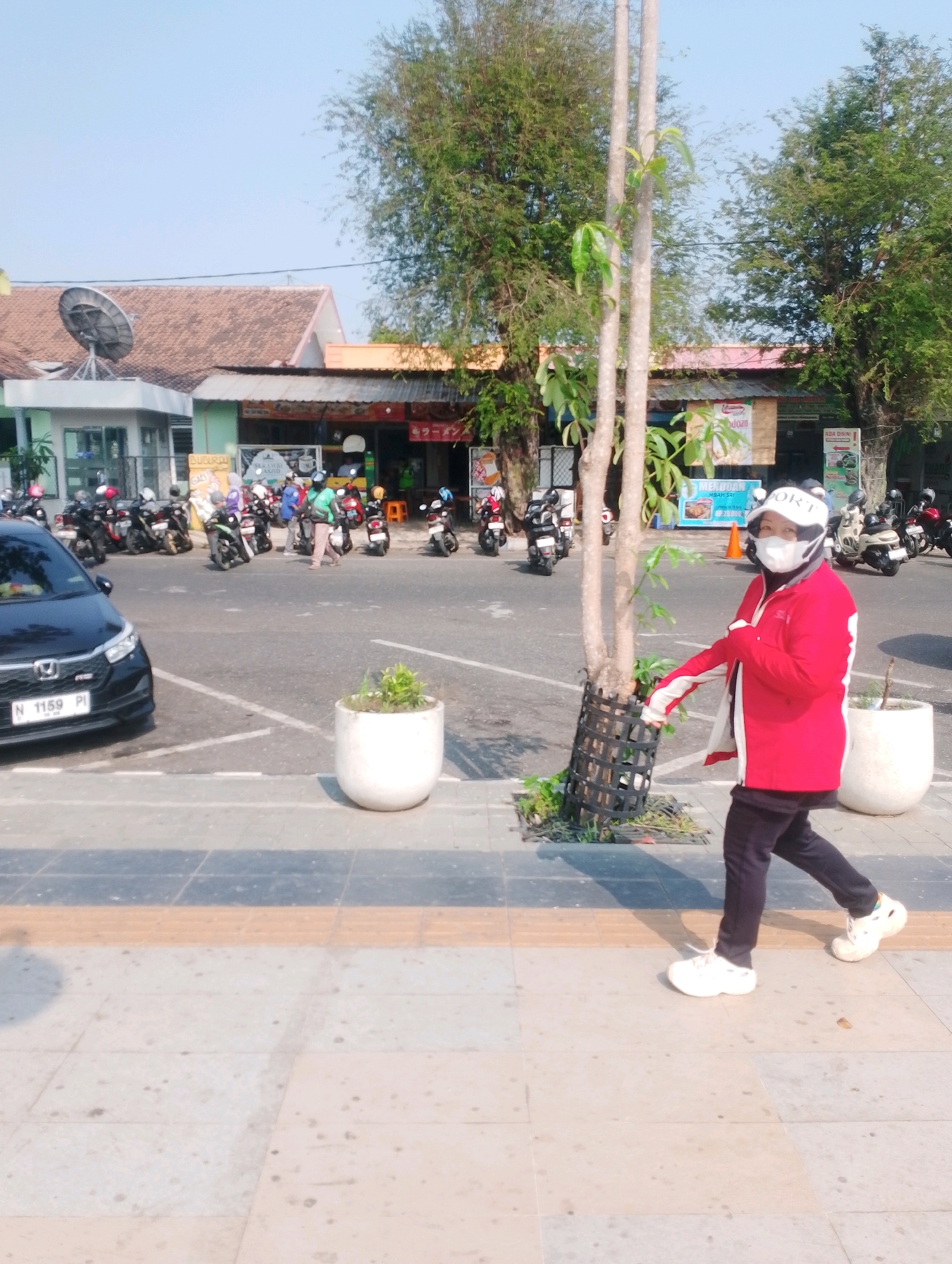 In Probolinggo, Indonesia, on a sunny day, a person wearing a red jacket, black pants, a white helmet, and a white face mask walks along a sidewalk next to a parking lot. The person appears to be a delivery driver. In the background, a row of shops and parked motorcycles are visible. The sky is clear and blue, and trees line the street. A black car is parked in the foreground.