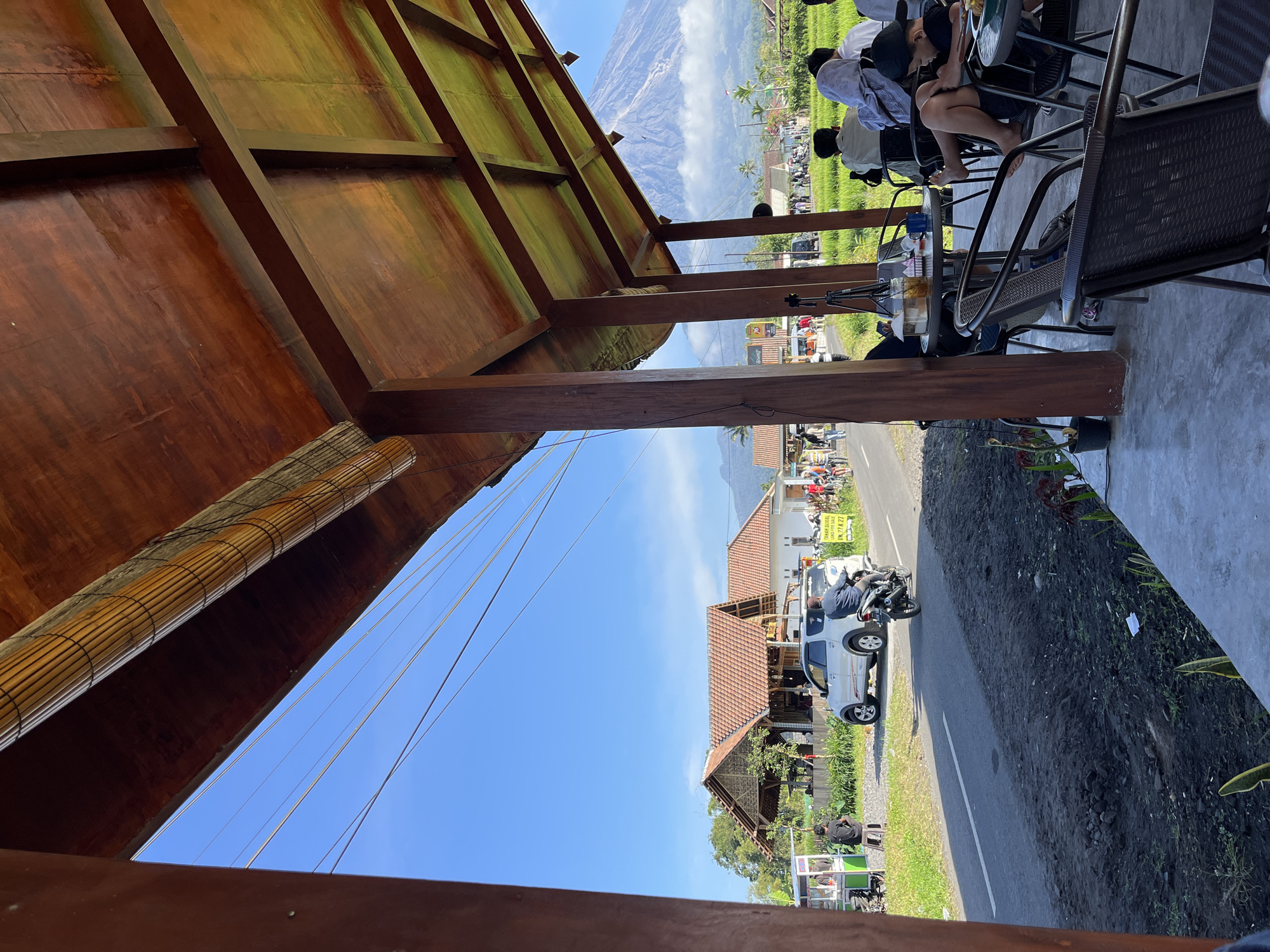 In the foreground, a covered patio with a wooden ceiling and pillars overlooks a scene in Oro-oro Ombo, Indonesia. Several people are seated at tables on the patio, some engaged with their devices or food. Beyond the patio, a paved road runs horizontally. On the left side of the road, there's a food cart and a white SUV. Further down the road, a motorcycle with a rider is visible, and beyond that, several buildings with red tile roofs and some people on bicycles. To the right of the road, lush green rice paddies stretch towards a mountain in the background, which appears to be Mount Merapi, a stratovolcano. The sky is clear and blue.