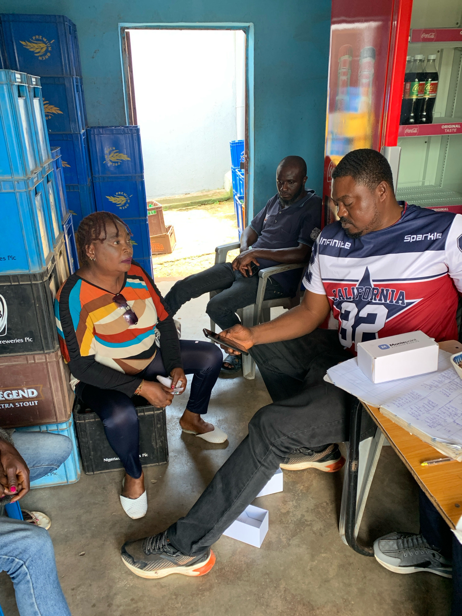 The image depicts an indoor scene, possibly a shop or storage area, in Jos, Nigeria. Three adults are visible, two men and one woman. The woman, seated on a black plastic crate, wears a colorful striped long-sleeved shirt and dark leggings. A pair of sunglasses is perched on her shirt, and she holds a small white object. She is looking to her right.
To her left, a man wearing a dark polo shirt is seated in a chair, looking down. To the woman's right, another man is seated in a chair at a table, wearing a blue, white, and red t-shirt with "CALIFORNIA 32" written on it. He is holding a smartphone in his right hand and looking at it. A white box with the "Moniepoint" logo is on the table in front of him, alongside a notebook with handwritten notes and a pen.
The environment features stacks of blue and brown plastic crates, some labeled with "PREMIUM BREW" and "LEGEND EXTRA STOUT," suggesting a beverage distribution or sales point. A red Coca-Cola refrigerator is visible on the right side of the image, stocked with bottles. The floor is concrete, and the walls are painted blue. An open doorway leads to an outdoor area with bright light, indicating daytime. The overall impression is that of a casual business transaction or meeting taking place in a somewhat cluttered, functional space.