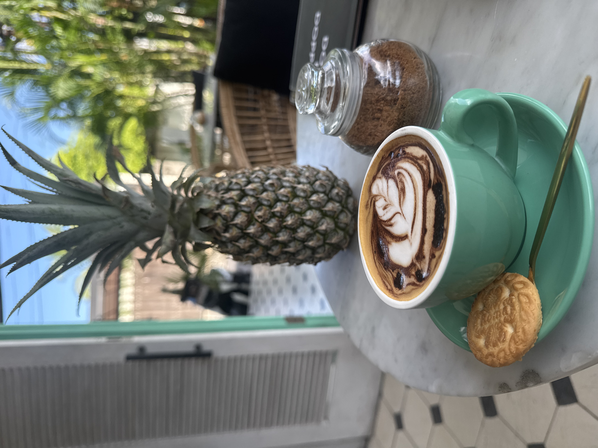 The image shows a table scene with a cup of coffee, a pineapple, and a jar of brown sugar. The coffee cup is light green with latte art on top and sits on a matching saucer with a small cookie and a gold-colored spoon resting on the edge. Next to the coffee is a jar of brown sugar and a whole pineapple. The table has a marble surface, and the floor has a tiled pattern of black and white squares. The location appears to be outdoors, possibly in a cafe, with trees and a building visible in the background. Based on the style and the objects present, this could be a setting in North Kuta, Indonesia. There are no people visible in the scene.