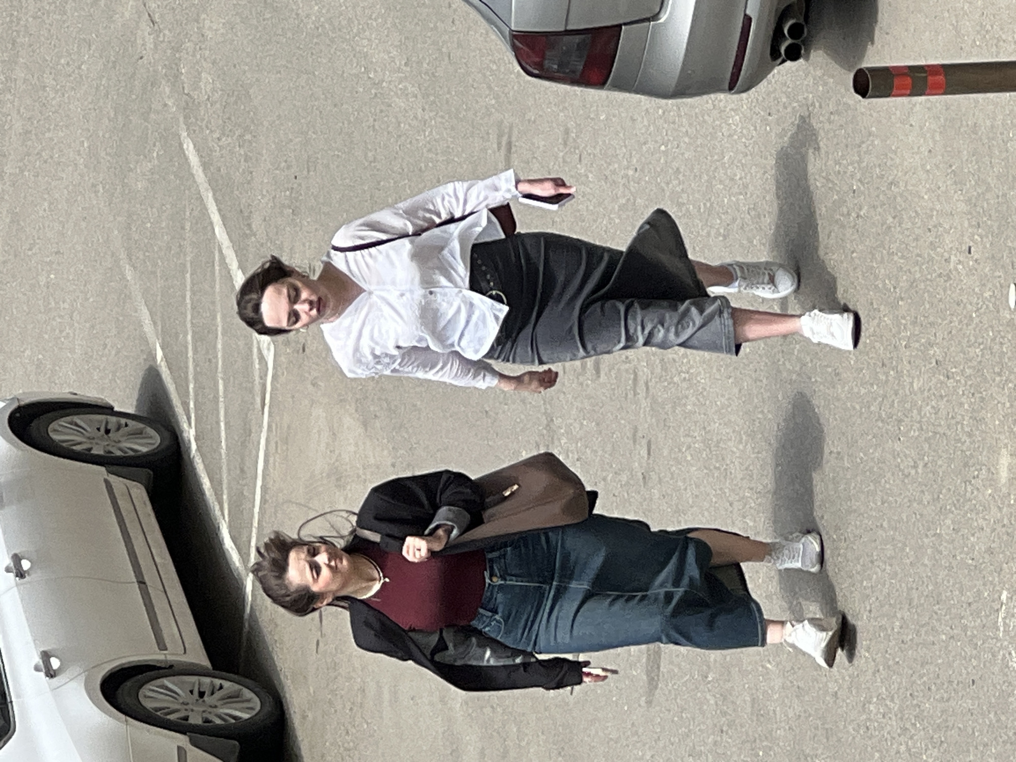 Two women are standing on a paved surface in what appears to be a parking lot. To the left, a white car is partially visible. Above the women, a silver car and a metal pole with red stripes are visible. The first woman wears a white shirt, a grey skirt and white sneakers and the second woman wears a dark red shirt, a denim skirt, a black jacket and white sneakers. Both women are holding objects in their hands. Based on the general appearance and style of the scene, it is likely to be a city in Russia, specifically Krasnodar.