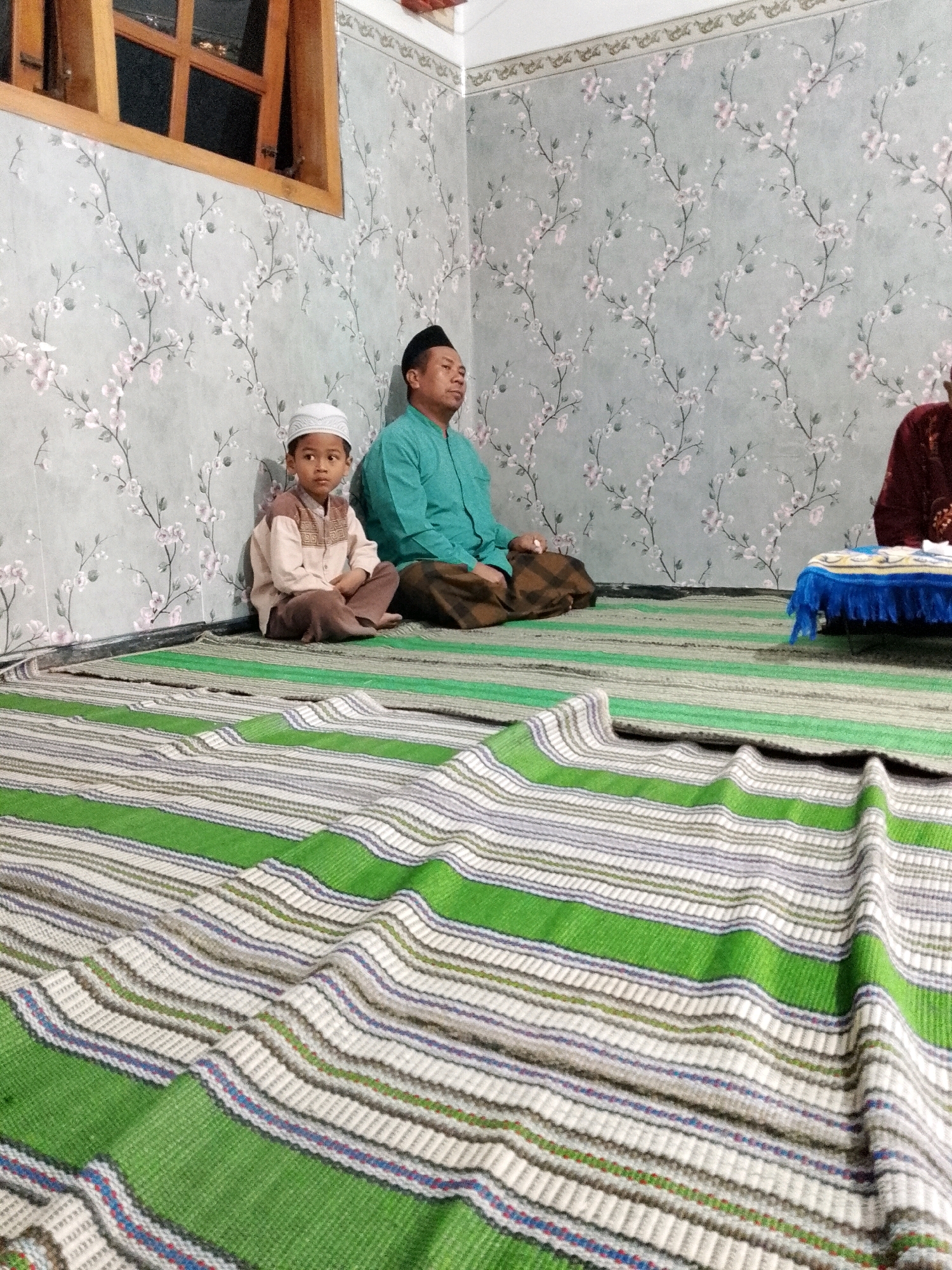 In Rejoyoso, Indonesia, an indoor scene depicts three individuals seated on a floor adorned with green and white striped rugs. A young boy in a white skullcap and a light brown and white tunic sits cross-legged in the foreground. To his right, a man wearing a teal collared shirt and a black skullcap is also seated. Further to the right, partially visible, is another person wearing a maroon garment. The room features walls with a floral wallpaper pattern in shades of grey and pink. A wooden window frame with multiple panes is visible on the upper left side of the image. A small, low table with a blue fringed cloth is placed near the person in the maroon garment. The overall lighting suggests an indoor setting, possibly during the day.