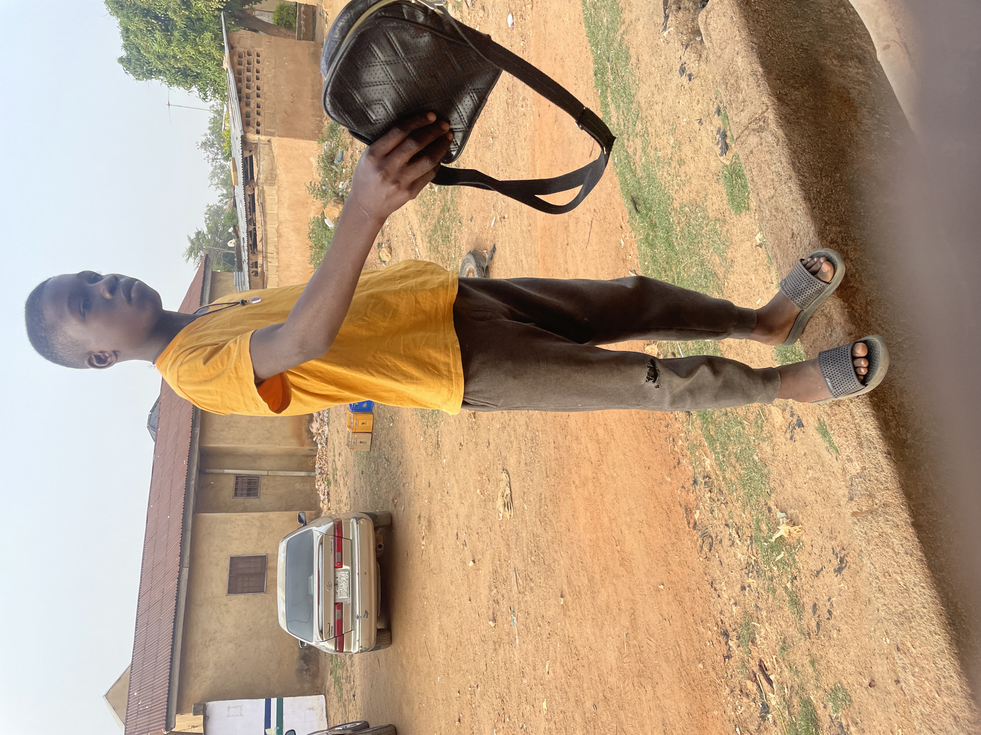 A young man stands on red dirt in Jos, Nigeria, holding a black quilted bag with a strap. He is wearing a yellow T-shirt, brown pants, and gray sandals. Behind him is a silver car, a tan building with a brown roof, and a tree. The ground is mostly dirt, with some patches of green grass.