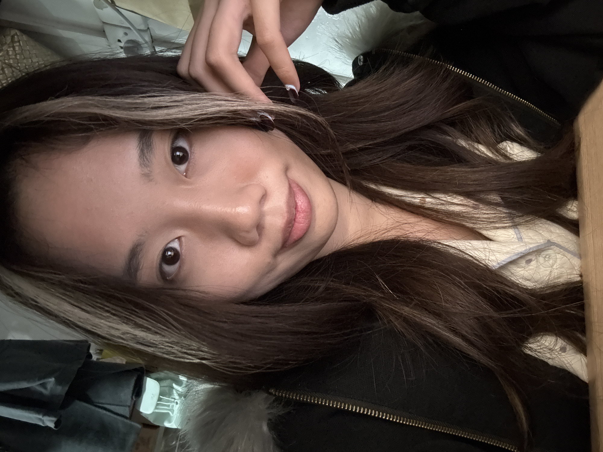The image displays a head-and-shoulders portrait of a young adult female, East Asian in appearance, positioned indoors. She possesses long, dark brown hair with distinct light blonde streaks framing her face. Her dark brown eyes are directed forward, and she exhibits a subtle, closed-mouth smile, revealing dimples on both cheeks.
The subject is wearing a black jacket with a visible gold-colored zipper, which is partially unzipped. A light-colored, possibly white or cream, textured garment is visible underneath the jacket. A light-colored, fur-like trim is present on the collar or hood of the jacket on the viewer's left. Her left hand is raised, with fingers (thumb, index, and middle) gently touching the side of her head near her right ear. The fingernails on these fingers are painted a dark color, with the index finger featuring a white tip.
The background indicates an indoor setting. To the upper left, dark gray fabric, possibly clothing, is hanging. Below this, a portion of a light teal or mint green backpack with white straps is visible. To the upper right, a white wall is present, on which a white electrical outlet with multiple sockets (consistent with Chinese Type I standards) is mounted, accompanied by visible wiring. Above the outlet, a light-colored, textured surface can be observed. The lower foreground consists of a light brown wooden surface, likely a table or desk. The electrical outlet type is consistent with infrastructure found in China, suggesting the image was captured within the country, potentially in the Nan'an District as specified.