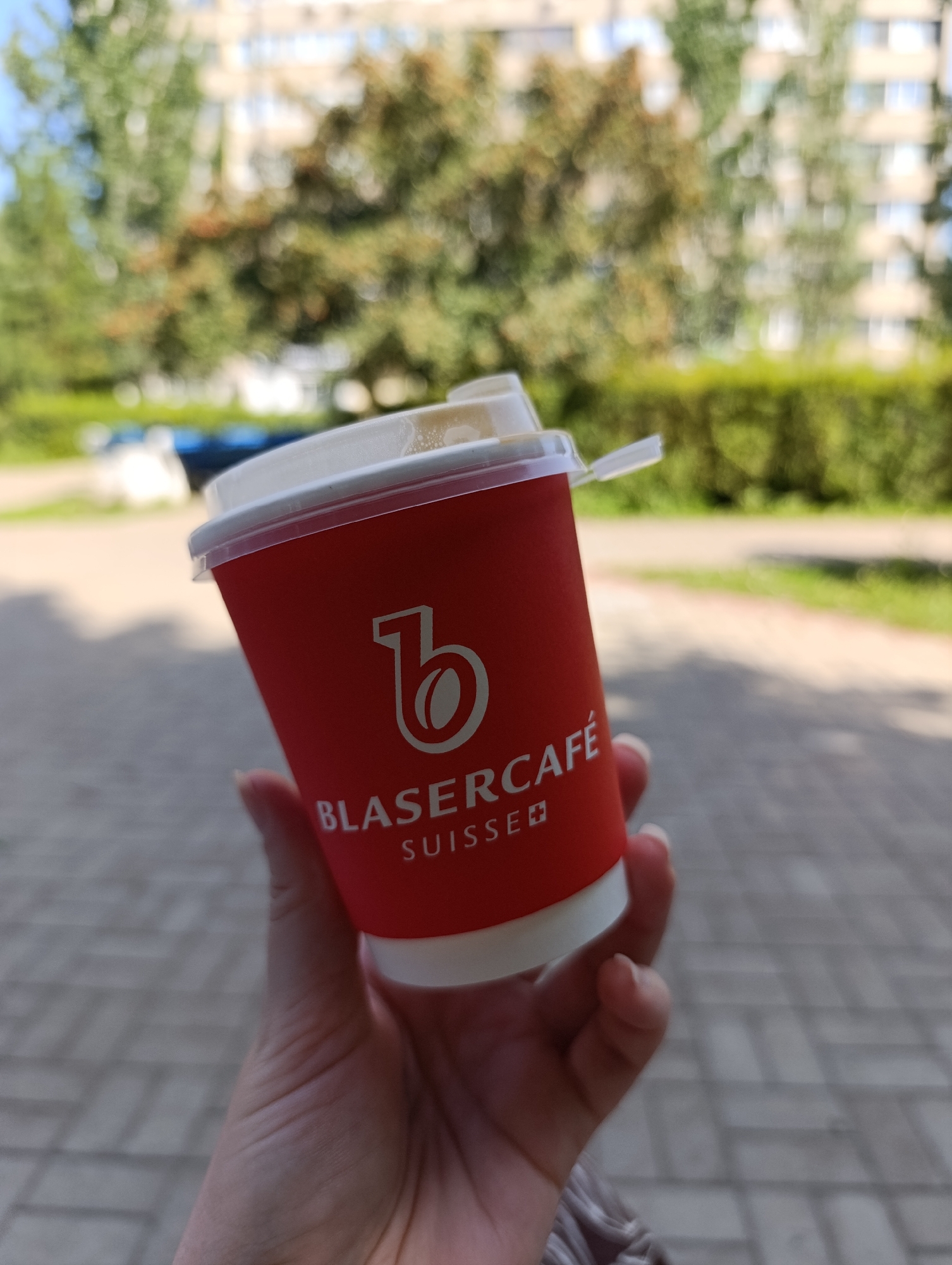 A person's hand holds a red disposable coffee cup with a white lid. The cup has a logo with the letter "b" inside a circle containing a coffee bean, and text below it reads "BLASERCAFÉ SUISSE". The background is blurred, showing trees and a building, consistent with an outdoor urban setting. The ground appears to be paved with bricks. This scene could be located in Tolyatti, Russia, where such coffee cups are common.