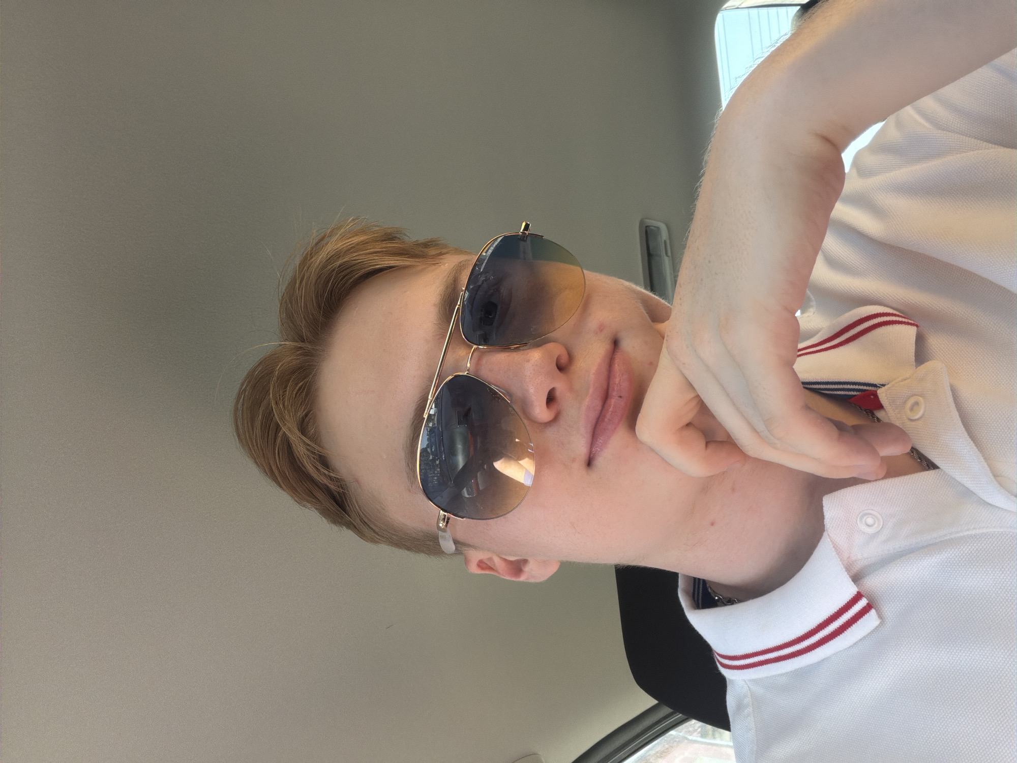 A young Caucasian male with blonde hair and fair skin is pictured indoors, possibly in a car, as indicated by the grey headliner and a dark seat visible. He is wearing aviator sunglasses with a gradient tint and a white polo shirt with red and blue trim on the collar. His right hand is resting on his chin, and he is looking at the camera. The background is blurred, with reflections of other vehicles visible in his sunglasses. The lighting suggests it is daytime.