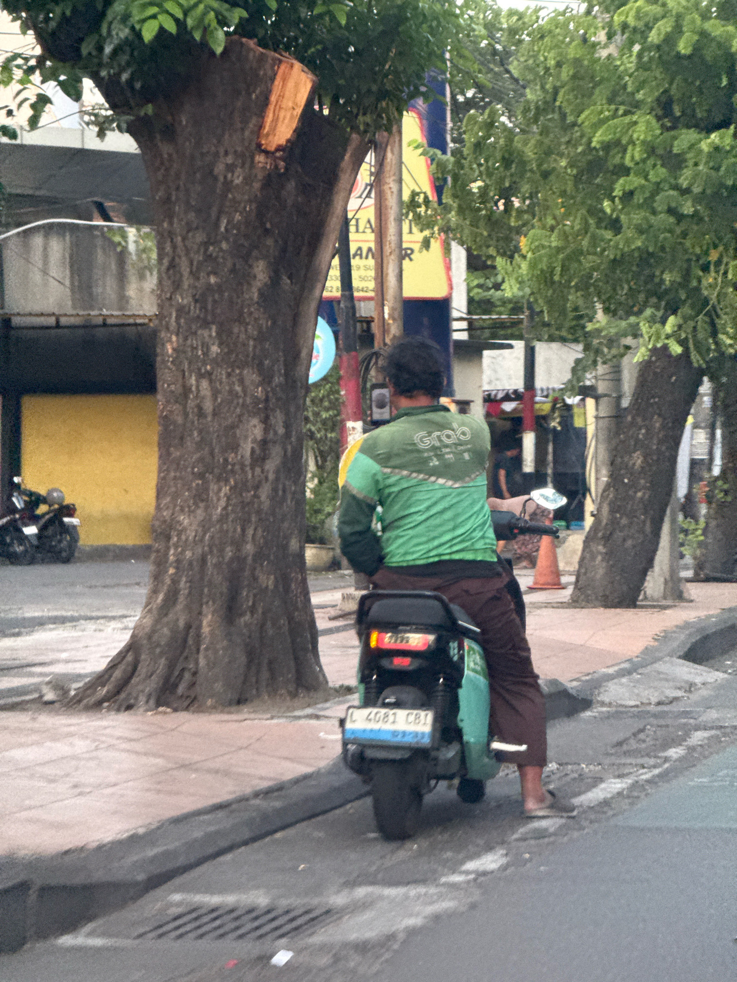A person in a green Grab uniform rides a motorcycle with a turquoise seat and black rear cargo box. The motorcycle's license plate is visible, displaying "L 4081 CB1". The rider wears brown pants and simple footwear. They are positioned on a street in Surabaya, Indonesia, indicated by the license plate, which starts with "L", a common prefix for vehicles registered in Surabaya and its surrounding areas. The environment features a large tree with a thick trunk to the left, a yellow wall in the background, and other trees and urban elements. The road surface appears worn, with visible patches and a sidewalk to the right. A few cones and traffic signs are also present in the background.