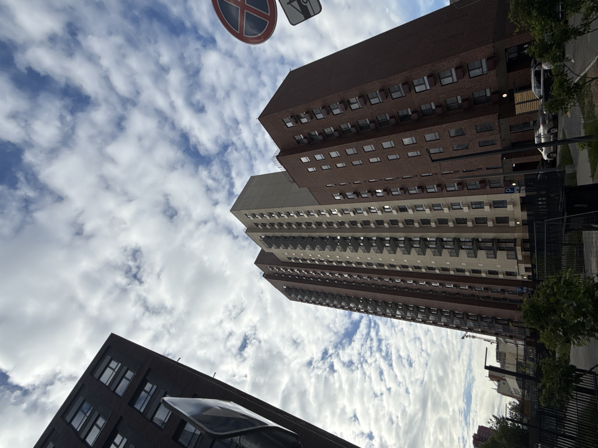 The image, taken from a low angle, captures several tall buildings against a backdrop of a cloudy sky. The buildings vary in color and architectural style. One building is primarily brown brick, another is a lighter beige color. The building at the bottom left is dark.
In the top left corner, there is a road sign. Vegetation can be seen on the right side of the image, along with a sidewalk, parts of other buildings, parked cars and a fence.
Given the architecture and style, it is plausible that this image was taken in Moscow, Russia.