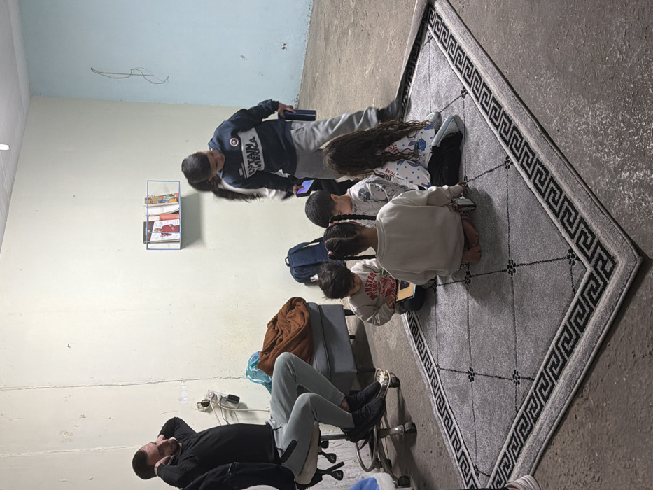 The image depicts an indoor scene within a functional, unadorned room, consistent with a community space or shelter in an urban setting like Tel-Aviv. The room features light beige walls, with a section on the right painted light blue, and a concrete floor. A grey rug with a black Greek key border and subtle central pattern is laid on the floor.
On the left, an adult male with a beard, wearing a black long-sleeved shirt, light grey sweatpants, and black sneakers, is seated on a silver rolling office chair. His hands are raised towards his head, appearing to touch his ears. Behind him, a grey ottoman is partially visible with a brown blanket draped over it, next to a blue backpack.
In the center and right foreground, six children are present. Five younger children are seated on the grey rug in a semicircle. Three are visible from behind, including a girl with pigtails wearing an off-white top and a girl with long dark hair in a patterned white top. Two other young boys are visible from the front-side, wearing long-sleeved tops, and appear to be engaged with small electronic devices (smartphones or tablets). One of the seated children is barefoot.
To the right, a slightly older boy stands, wearing a dark blue "Captain America" hoodie, light grey pants, and dark shoes. He holds a smartphone in his left hand and a dark blue bottle or container in his right. Another individual is partially visible behind him.
A small white wall-mounted shelf containing a few books or magazines, one featuring a person in red attire, is present on the back wall. Electrical outlets and minor wall imperfections are visible on the beige wall. The scene captures a moment of adults and children occupying a shared space.