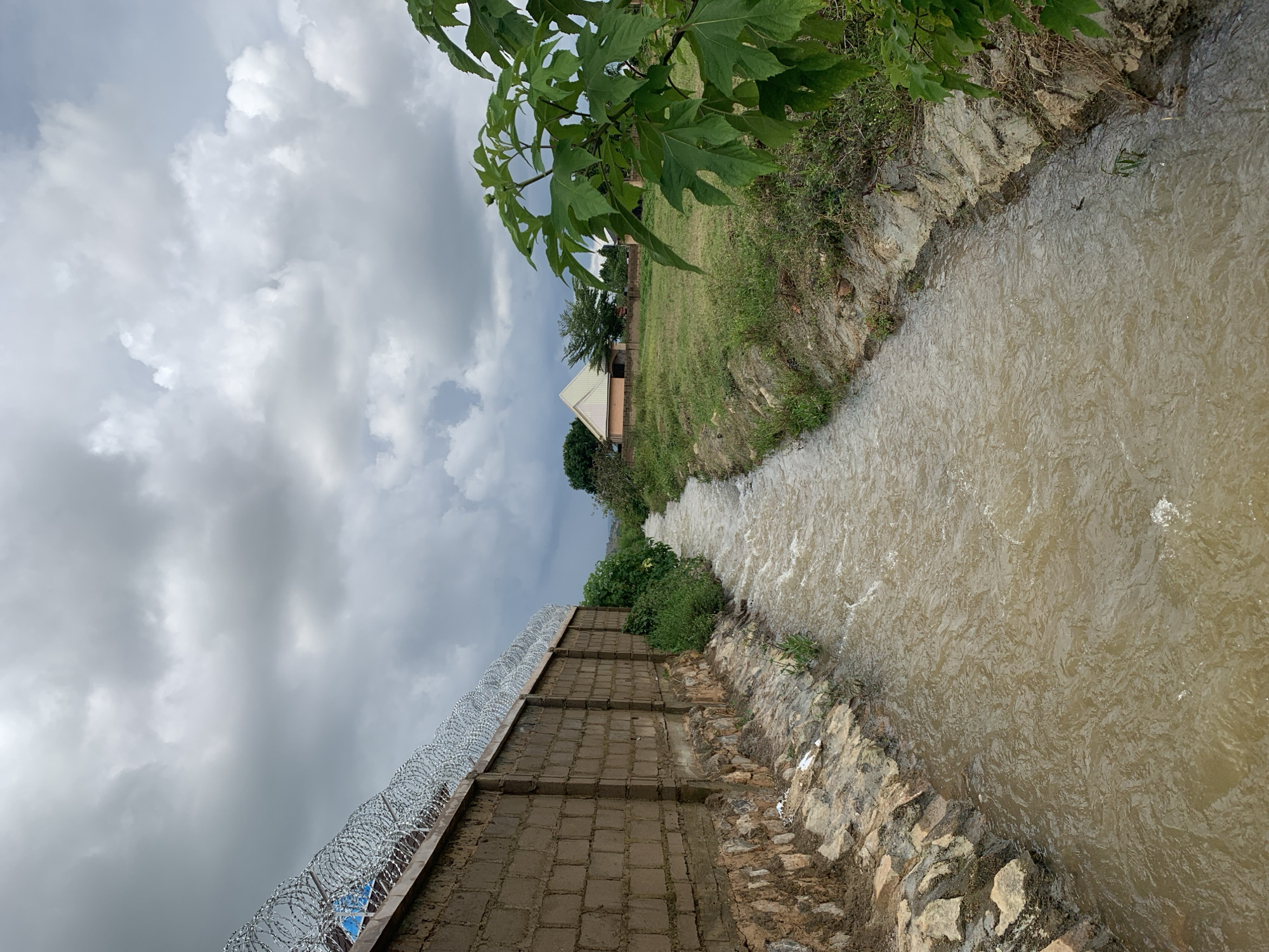 The image shows a channel of muddy water flowing between two banks. The left bank is lined with a brick wall topped with razor wire. The right bank is covered with grass and vegetation, with a large green plant in the foreground. In the background, there is a grassy field with a light-colored building with a triangular roof. The sky is overcast with dark clouds. This scene is located in Gura Top, Nigeria.