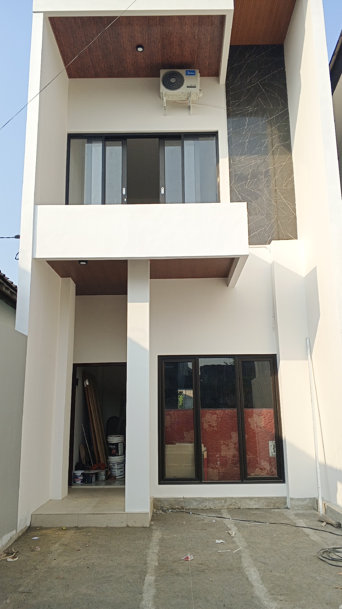 The image shows the exterior of a modern, multi-story house with a clean, white facade. The house features large, dark-framed windows. On the upper level, there is a balcony with a wooden-grained ceiling and an air conditioning unit mounted on the wall. A section of the upper facade is decorated with dark, marbled tiles.
The ground floor has an open entryway where several buckets, likely containing paint, and long wooden planks are stored. Adjacent to the entryway is a large window with red patterned material visible behind the glass. The ground in front of the house is a paved area with visible concrete and some scattered debris. The lighting suggests it is daytime, with sunlight casting shadows. The location is identified as Ambarketawang, Indonesia.