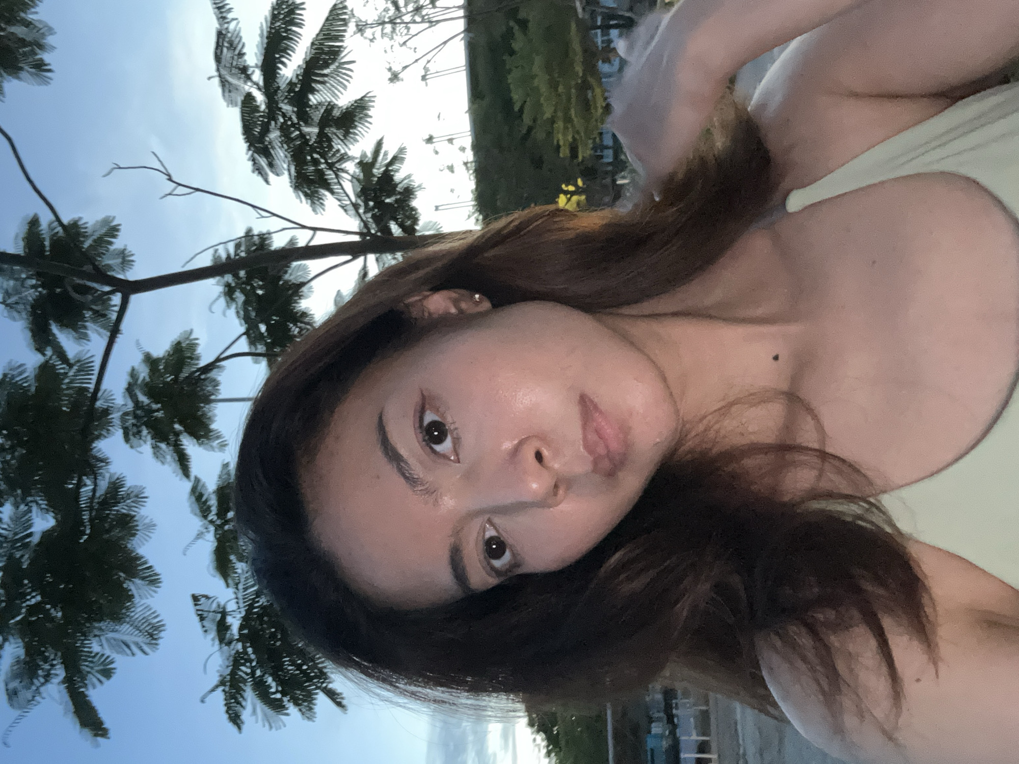 A medium shot selfie shows a young woman with long, dark brown hair and fair skin. She is wearing a light green top. Her gaze is directed slightly to her left, and her expression is neutral. She has a small mole on her neck. Behind her, palm trees and a blue sky are visible, suggesting an outdoor setting. The lighting indicates it might be late afternoon or early evening. The background includes hints of buildings and greenery, suggesting a populated area.