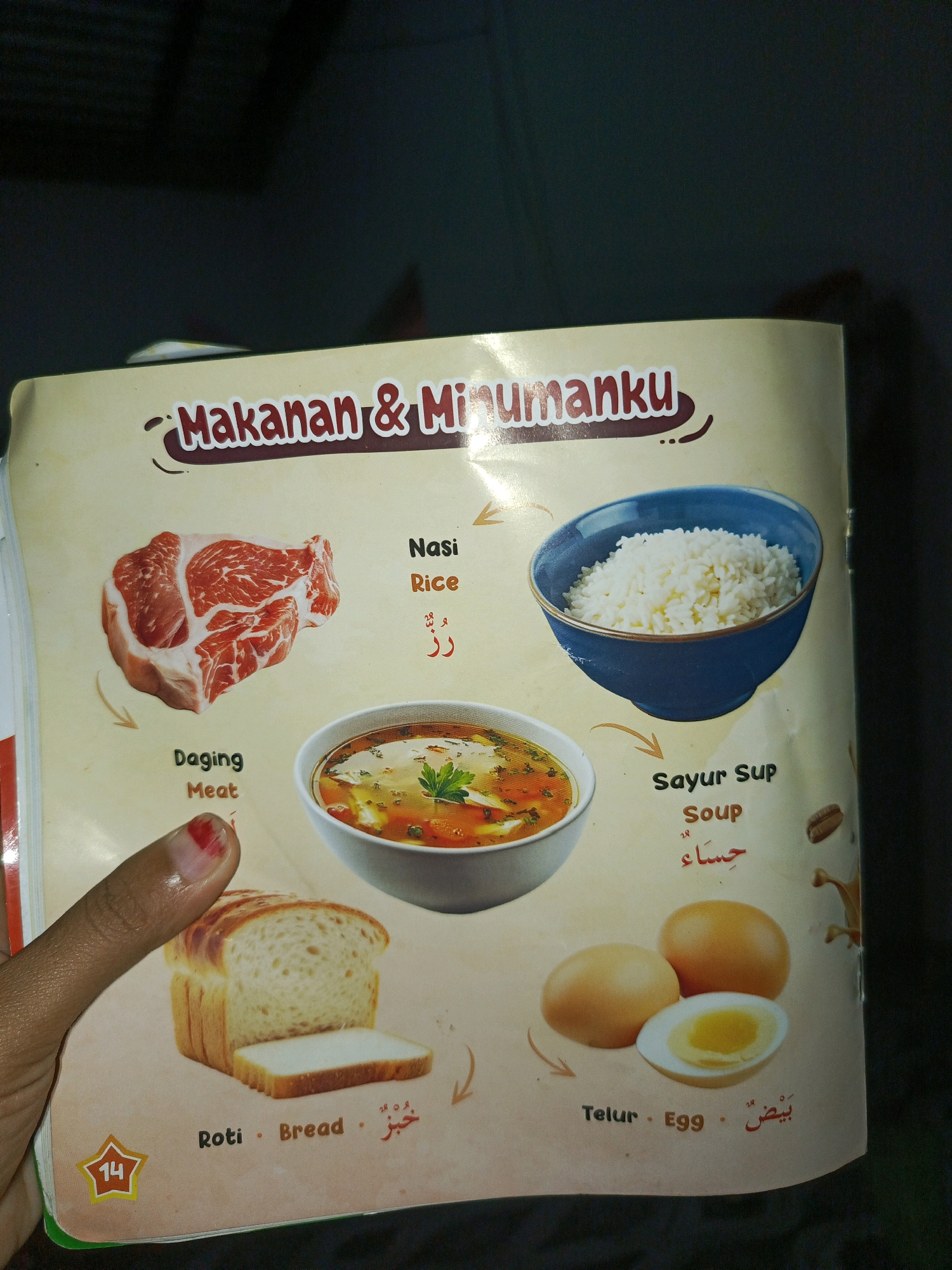 The image is a close-up shot of a page from a children's book or learning material, featuring various food items with their names in Indonesian and English. The background is a light peach color.
In the top center, the title "Makanan & Minumanku" is displayed in stylized red lettering.
To the left, there is a raw piece of red meat, labeled "Daging" and "Meat".
To the right of the meat, a blue bowl contains white rice, labeled "Nasi" and "Rice".
Below the rice, a white bowl contains a soup with vegetables and what appears to be chicken or meat, labeled "Sayur Sup" and "Soup".
In the bottom left, a loaf of bread is shown, with one slice removed, labeled "Roti" and "Bread".
To the right of the bread, three eggs are depicted, one of which is halved to show the yolk and white, labeled "Telur" and "Egg".
A yellow star with the number 14 inside is visible in the bottom left corner.
A finger with red nail polish on the thumb is partially visible in the bottom left, pointing towards the bread.
There are no discernible features or elements in the image that would indicate a specific location in the city of Megang Sakti I, Indonesia.