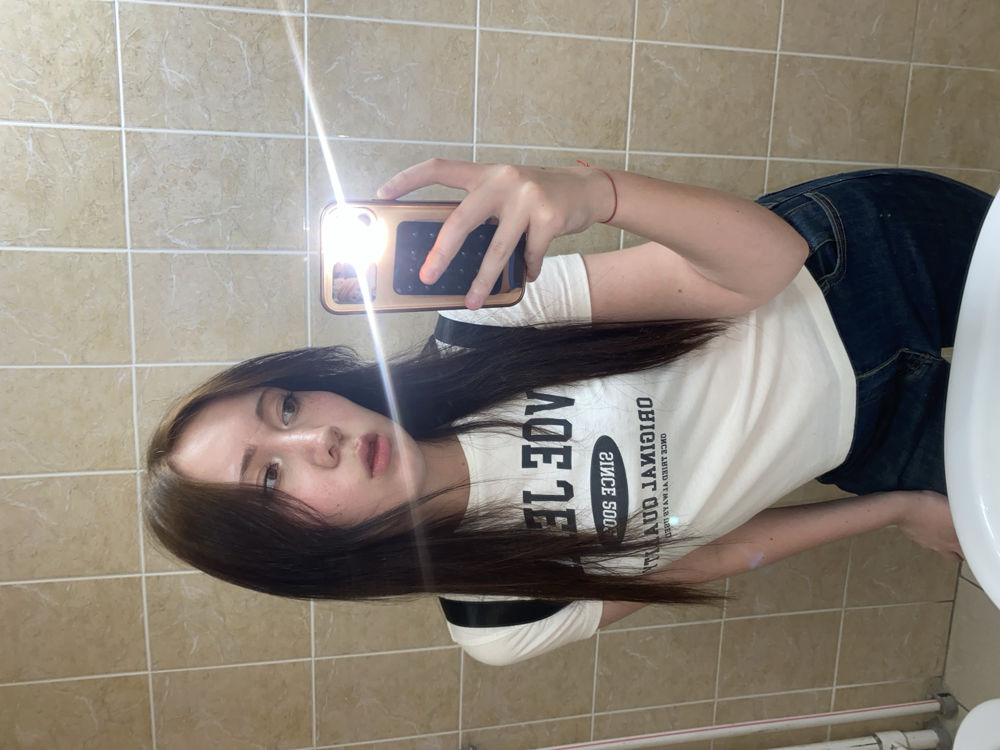 A person is taking a selfie in what appears to be a bathroom with beige tiles. The person is wearing a white t-shirt, dark jeans, and has long brown hair. The phone has a gold case and the flash is on. The location appears to be indoors, possibly a private residence or a public restroom.