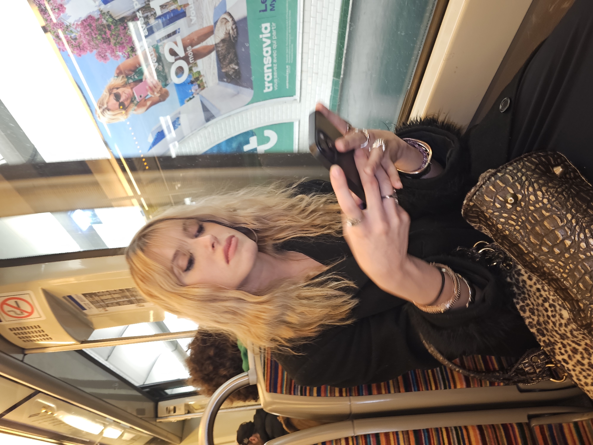 The image shows a woman with blond hair, wearing a black jacket, sitting on public transportation, possibly a bus or a metro in the city of Paris, France. She is looking at her smartphone and has multiple rings on her fingers and bracelets on her wrist. Next to her is a brown patterned handbag. The seat has striped upholstery. In the background, through the window, there is an advertisement featuring a blonde woman taking a selfie and the logo of "transavia," a French airline. The seats and ceiling indicate that the picture was taken inside a bus.
