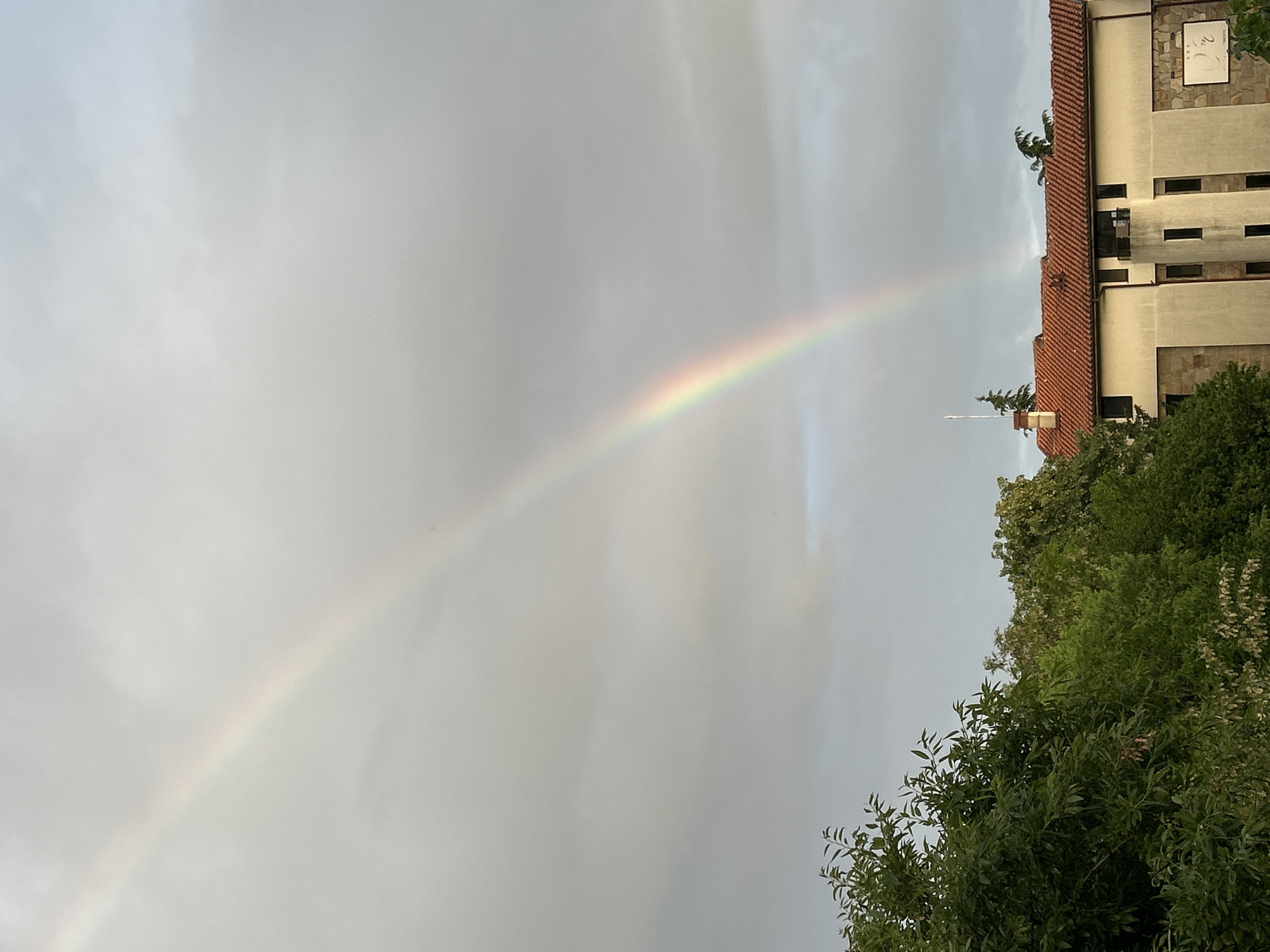 The image shows a rainbow arching across a cloudy sky. The right side of the image includes the top of a building with a red tile roof, light-colored walls, and dark window openings. Trees are visible in the lower right corner, partially obscuring the base of the building. A sundial is visible on the building near the roofline. The location is Polenitsa, Bulgaria.