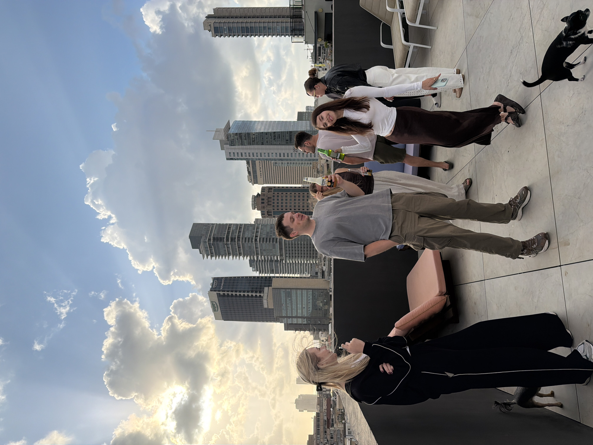 The image depicts a gathering of people on a rooftop in Tel Aviv, Israel. The background features a cityscape with skyscrapers and a partly cloudy sky. Several individuals are standing or sitting, some holding drinks, and there's a dog present. The setting appears to be a casual social event.