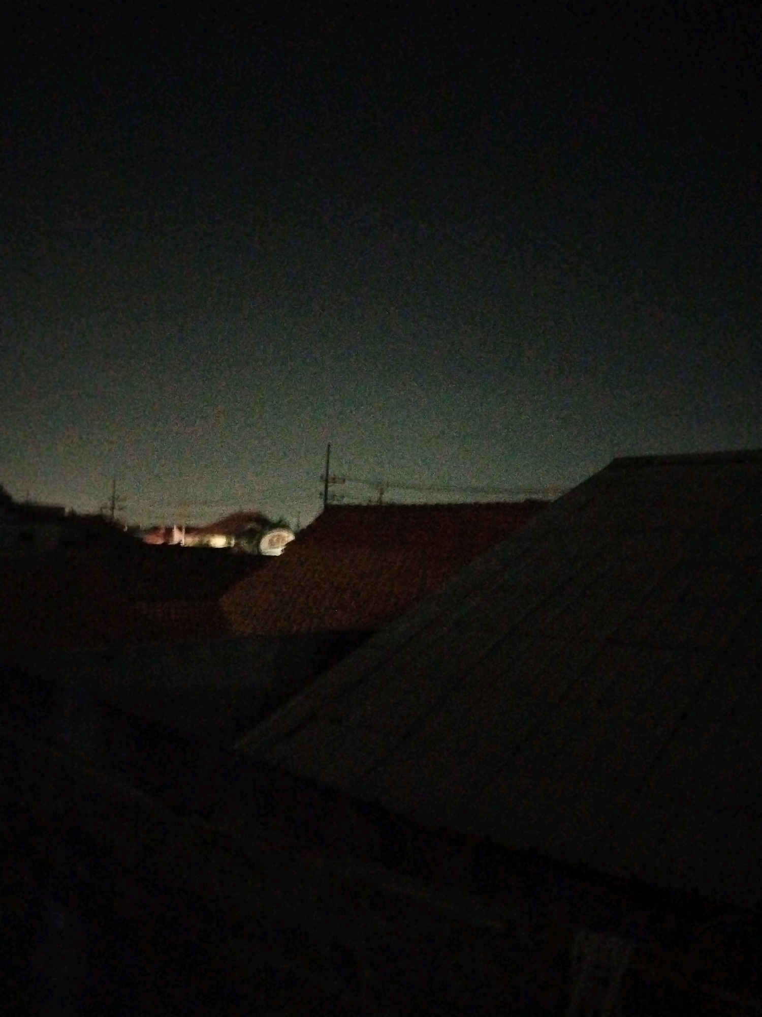 The image captures a nighttime scene overlooking rooftops in Pepelegi, Indonesia. The sky is dark, with some distant lights suggesting a populated area. Several rooftops are visible, some with a reddish hue, possibly from terracotta tiles, while others appear darker. A few faint vertical structures, likely utility poles, are interspersed among the buildings. In the distance, there are scattered illuminated buildings and a circular object. The overall impression is one of a residential area at night, with a sense of quiet and distance. There are no people or discernible actions in the frame.