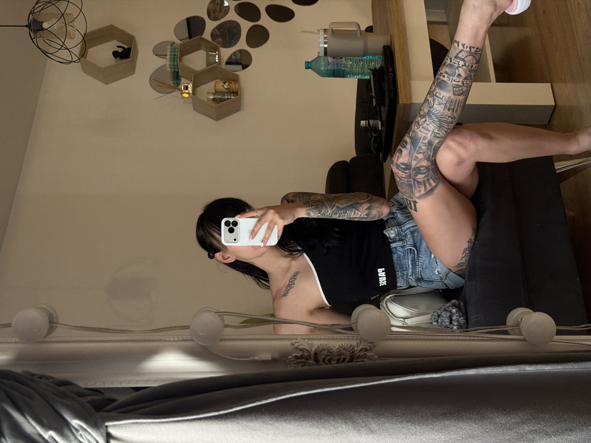 The image shows a young woman with dark hair, wearing a black one-shoulder top and denim shorts, sitting on a dark gray sofa. She is holding a white smartphone in her right hand and taking a selfie in a mirror. The woman has extensive tattoos on her arms and legs.
The background suggests an indoor setting, likely a living room. There is a wooden table in the foreground with a blue water bottle, a large insulated mug with a straw, and other items. Behind the table, a dark sofa is visible. In the upper right corner, there are hexagonal shelves on the wall, decorated with various objects, including small figurines and mirrors. A contemporary pendant light fixture hangs from the ceiling. To the left, a grey curtain and a string of decorative lights are visible, reflecting in the mirror.
The image does not contain any information or visual cues that would allow for the identification of the specific city or location as Plovdiv, Bulgaria.
