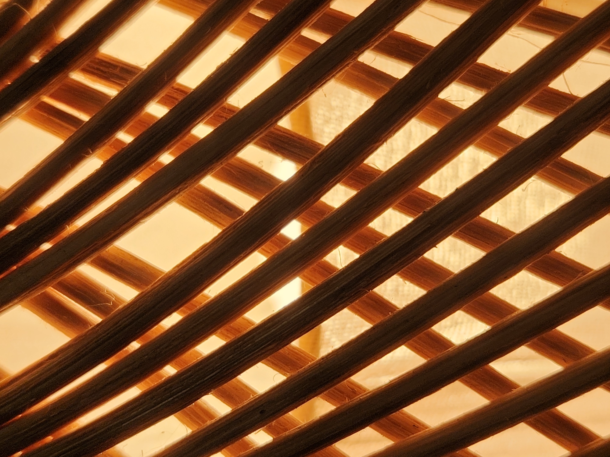 This image is a close-up, abstract photograph of a woven pattern. The primary subjects are the diagonal strands of woven material, appearing to be natural fibers such as rattan or bamboo, with a darker, more substantial layer of strands forming the foreground and a lighter, thinner layer creating the background. The material is illuminated from behind, casting a warm, golden-orange glow through the gaps in the weave. This backlighting creates a strong contrast between the dark, textured foreground strands and the bright, diffused background.
There are no people, animals, or specific objects clearly identifiable. The scene is abstract and textural, focusing on the interplay of light, shadow, and the woven pattern. The texture of the material is highly detailed, showing the grain and fibers of the strands. There are no clear cues about the time of day, weather, or specific location beyond what can be inferred from the material itself. The overall mood is warm and somewhat tranquil, due to the soft, diffused light. No text is visible in the image. The context provided suggests Maitland, United States of America, but this cannot be confirmed from the visual information alone.