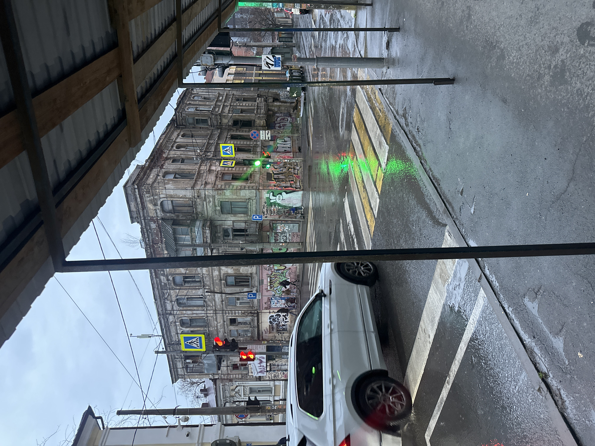 The photograph, taken in Rostov-on-Don, Russia, shows a wet street scene viewed from under a covered walkway. An old, multi-story building with graffiti and peeling paint stands across the street. Multiple traffic signs and signals are visible, some showing red lights and others green. A white car is stopped in a crosswalk, partially obscuring the view of the building. The street is wet, reflecting the traffic lights and sky. The sky is overcast.
