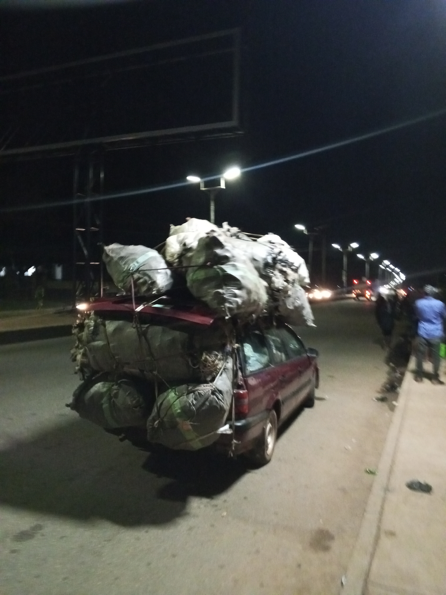 A photograph taken at night on a road in Bukuru, Nigeria, depicts a burgundy station wagon overloaded with large bags. The bags are stacked on the roof of the car and secured with ropes. Some bags are also tied to the back of the vehicle. The scene is illuminated by streetlights. A few people are visible on the sidewalk, and other vehicles are on the road in the distance. The road surface appears to be asphalt or concrete.