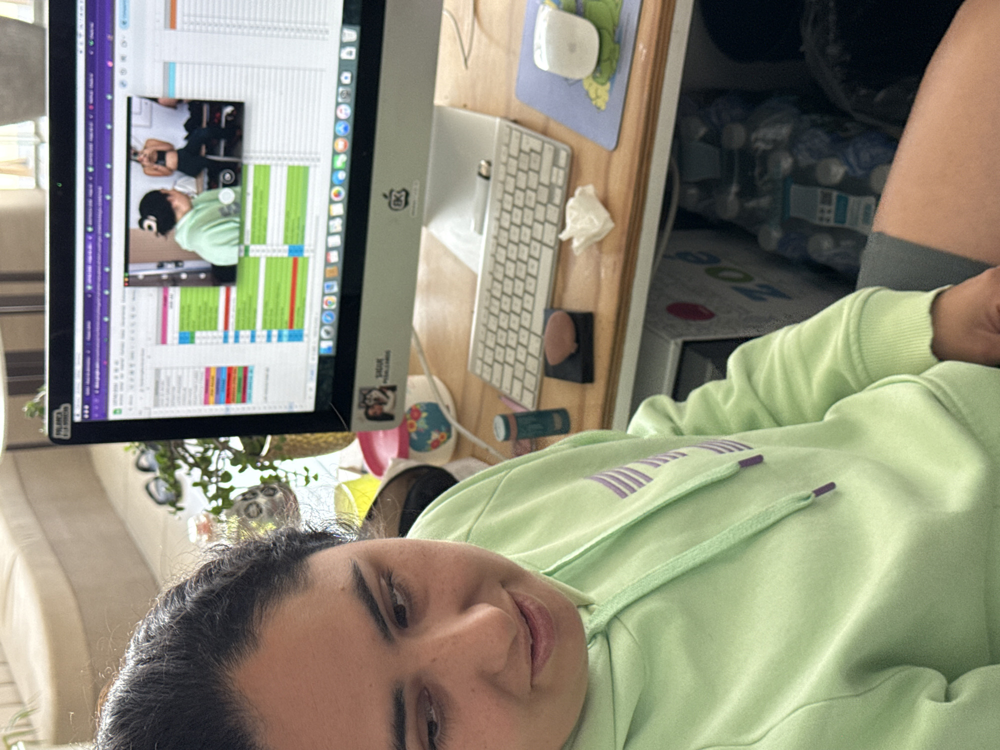 A woman is lying down, facing the camera. She is wearing a light green hoodie with purple stripes on the hood. Behind her is a computer monitor displaying a spreadsheet, and a video call showing another woman exercising. Next to the monitor is a keyboard, mouse, mousepad, and other small objects on a wooden desk. A pack of water bottles is visible behind the desk. There is no information in the image that explicitly indicates the location is in the city of Puebla, Mexico.