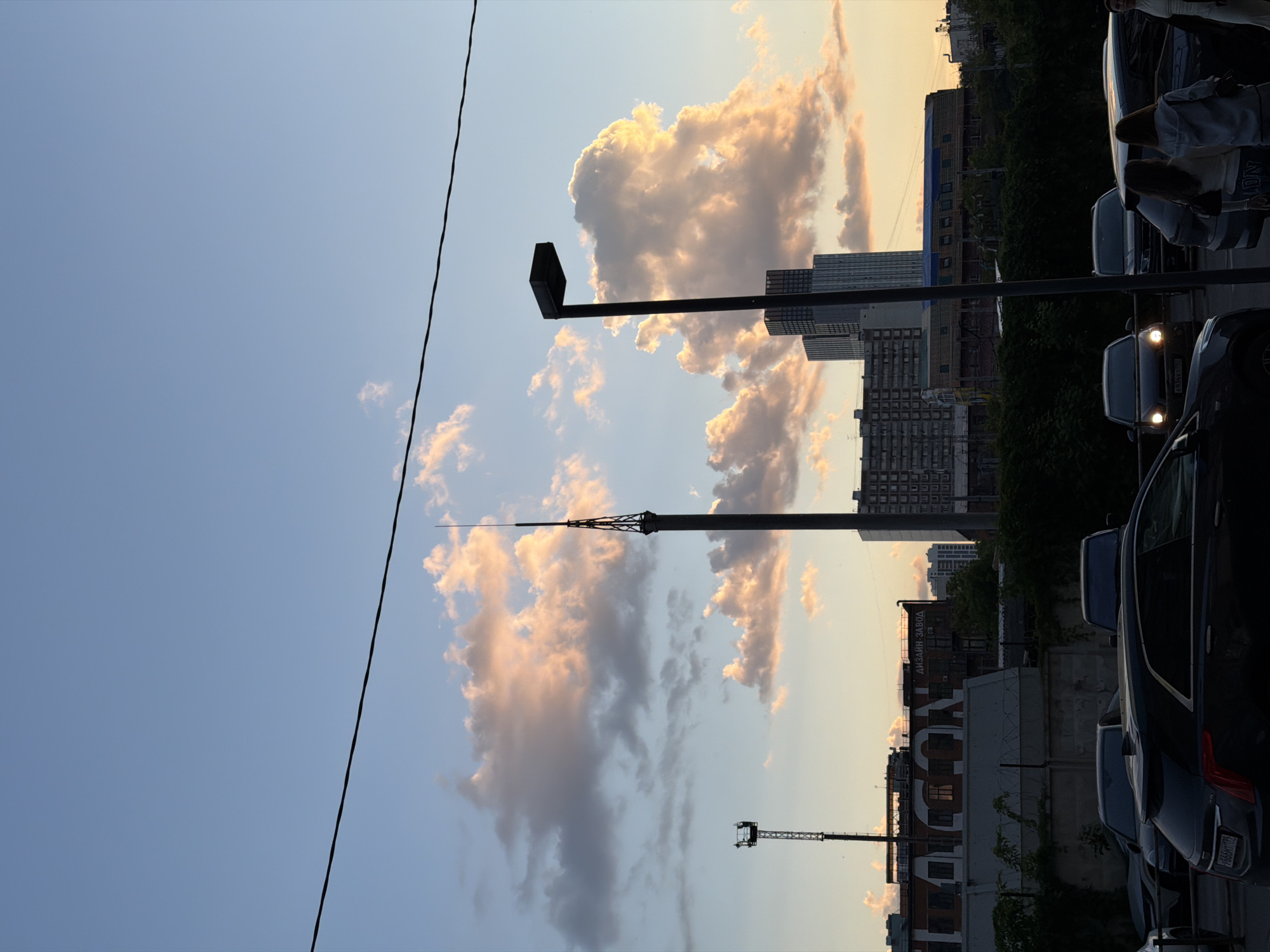 The image depicts an urban street scene captured during the golden hour, with the sun setting behind the buildings. The sky transitions from clear blue at the top to warm yellow and orange hues near the horizon, illuminated by scattered, brightly lit cumulus clouds displaying golden and pink tones.
A prominent, slender communications or TV tower with a spired top stands tall in the midground, silhouetted against the vibrant clouds. To its right, a tall, dark streetlight with a rectangular fixture is also silhouetted. A thinner, diagonal power line stretches across the upper left portion of the sky.
Below the horizon, a cityscape unfolds with various buildings, including multi-story structures and industrial facilities. A notable industrial-style building in the midground features a large white sign with visible Cyrillic text: "ДИЗАЙН-ЗАВОД" (DESIGN FACTORY). Below this, another word, possibly "FLACON", is partially visible. Dark vegetation, possibly trees and shrubs, is visible between some buildings and the street level.
On the right side of the image, several cars are parked along what appears to be a street or parking area. Some vehicles have their headlights illuminated. Indistinct silhouettes of a few people are visible near the cars, with one person standing near the far right edge. A license plate on one of the cars is visible, reading "Y766OO 199RUS". The scene evokes a calm, everyday atmosphere in an urban setting, characteristic of late afternoon or early evening in Khimki, Russia.