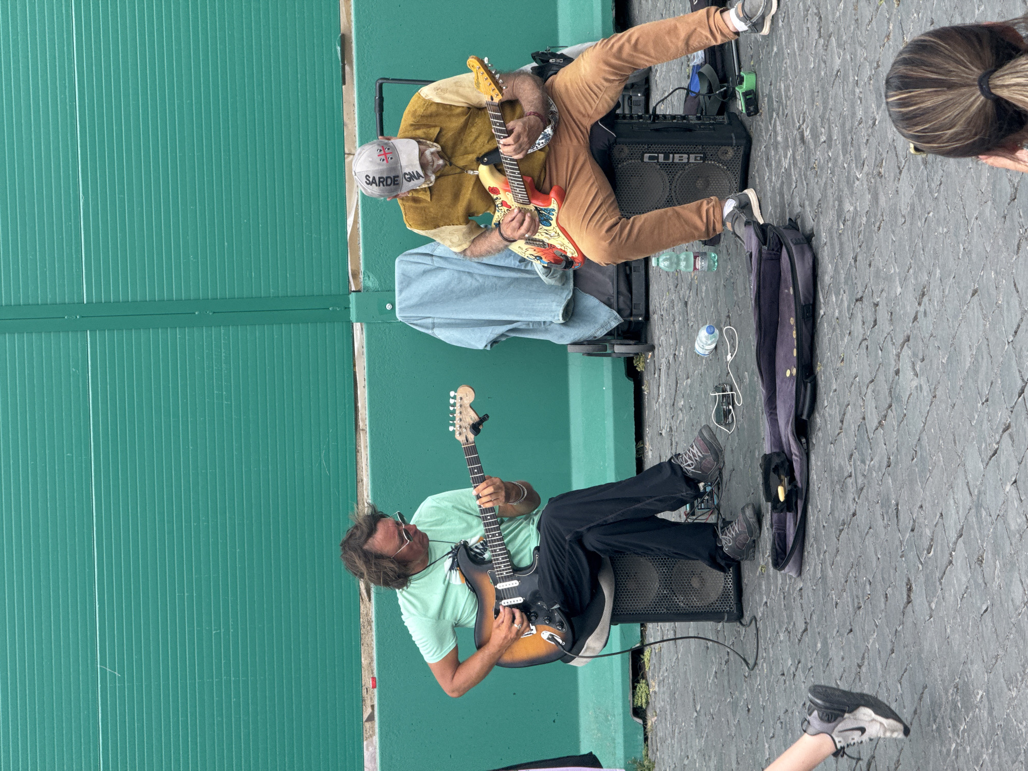 The image captures a street performance by two male guitarists against a teal-green corrugated metal wall, likely in Rome, Italy. The ground is paved with grey cobblestones.
In the upper part of the frame, one musician is seated, playing a red electric guitar with a colorful, intricate design (possibly floral or patterned). He wears a light brown button-up shirt, brown trousers, and a white cap with "SARDEGNA" and a flag-like emblem (resembling a Union Jack cross) visible on the front. He has a beard. His leg rests on a black "CUBE" brand guitar amplifier. A light blue fabric covers a surface beneath him, possibly a stool or case.
In the lower part of the frame, a second musician is also seated, playing a sunburst-finish electric guitar (resembling a Fender Stratocaster). He wears a light green t-shirt, dark trousers, and sunglasses. His hair is long and dark. He is seated on a black guitar amplifier. Next to him, a dark purple guitar case lies open on the cobblestones.
Various plastic water bottles are scattered on the ground near both musicians' equipment. A person's head with brown, streaked hair tied back is partially visible on the far right, suggesting an audience member. The scene appears to take place during the day, under ambient light.