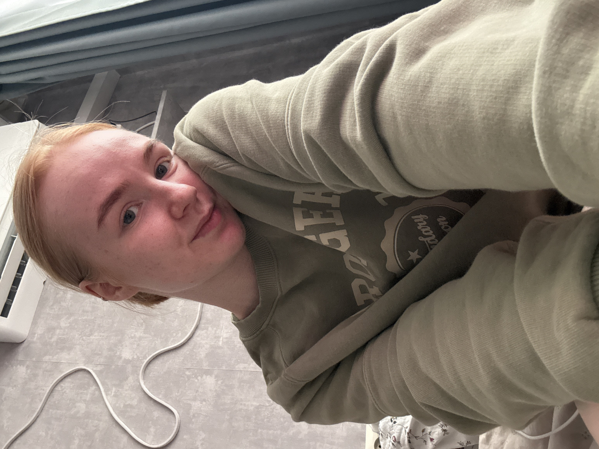 A close-up, angled selfie of a fair-skinned individual with light blonde hair, wearing an olive green sweatshirt with a printed design on the chest. The person is looking directly at the camera. The background consists of a grey floor with white electrical cords, a white radiator, and light blue curtains. There is no visible information to suggest the location is Chaikovsky, Russia.