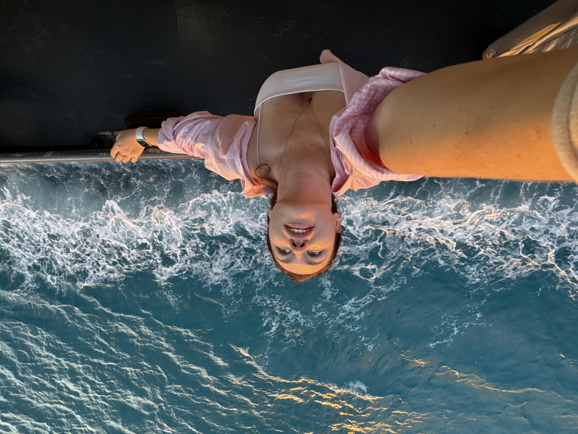 An overhead medium shot captures a woman taking a selfie as she leans over the railing of a boat, looking up and to her left with a slight smile. She is wearing a pink and white striped open shirt over a white tank top, a necklace with a pendant, and a white smartwatch. Her left arm is extended, holding the camera, and her right hand rests on the boat's dark railing. The choppy, turquoise ocean with white foam stretches out behind her, illuminated by golden light, suggesting either sunrise or sunset. The scene is framed from a high vantage point, emphasizing the vastness of the water.