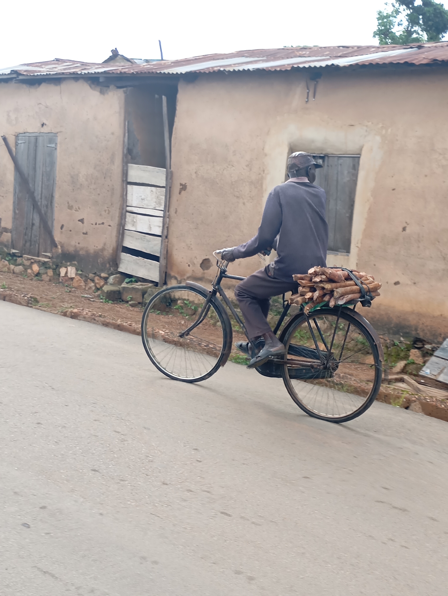 A man rides a bicycle loaded with firewood down a paved road in Shendam, Nigeria. The man, wearing a dark long-sleeved shirt and dark pants, is seen from behind, with a cap on his head. The bicycle is black with a rack on the back carrying a bundle of firewood tied with string. The road is gray asphalt. In the background, there is a building with a corrugated metal roof and a mud or stucco exterior. The building has two wooden doors and appears to be a residential or commercial structure. The ground next to the road is dirt with some scattered stones and vegetation. The sky is overcast.