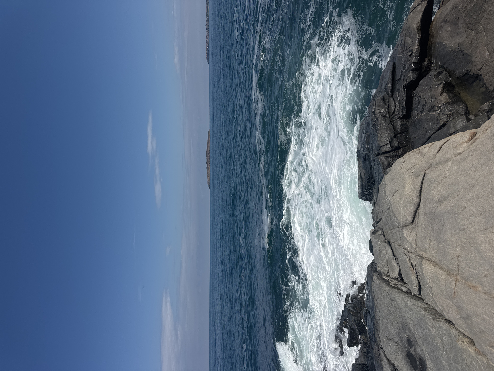 The image depicts a rocky coastline with the Black Sea in the background. In the foreground, large, grey rocks are prominently featured, with dark, wet rocks closer to the water's edge. Waves are crashing against the rocks, creating white foam and spray. The sea is a deep blue, with ripples and choppiness. In the distance, a small island with vegetation is visible, and further to the right, a town with buildings can be seen along the coast. The sky is a clear, vibrant blue with a few wispy white clouds. The scene appears to be a natural coastal landscape, likely taken during daylight hours.