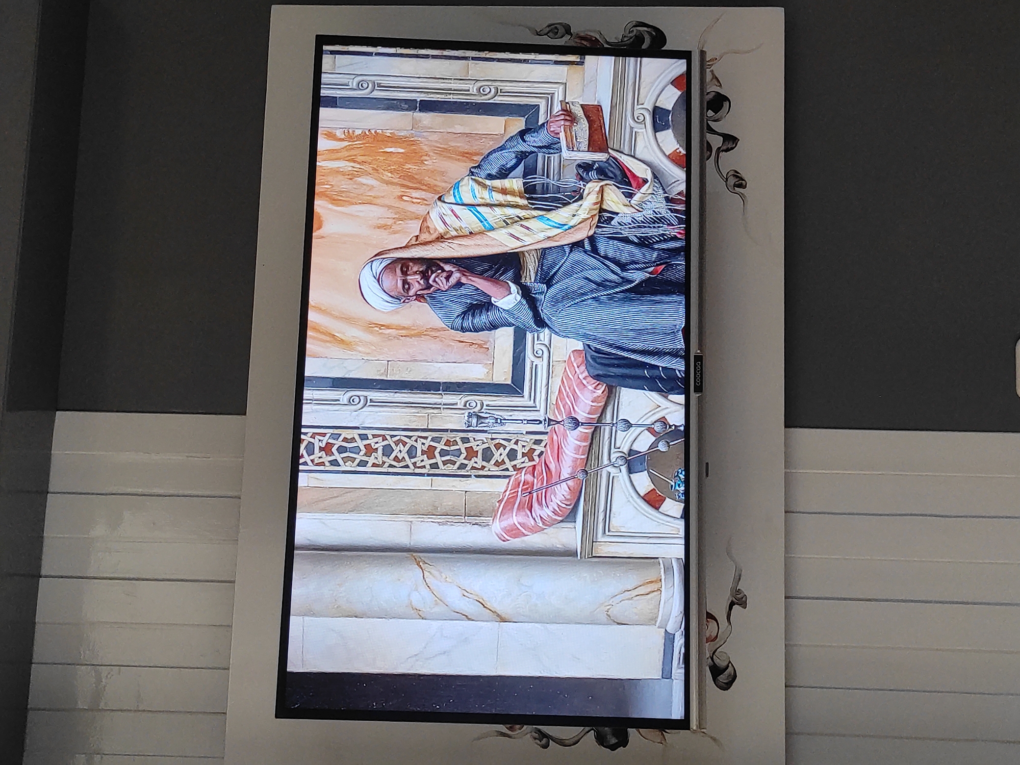 The image shows a television screen displaying a painting of a man in traditional Middle Eastern attire. The man is seated on a bench, resting his chin on his hand, and appears to be deep in thought. He wears a white turban, a striped robe, and a colorful patterned shawl draped over his shoulders. Beside him is a small wooden box. The background of the painting features architectural elements with intricate patterns and marble columns. The television itself is mounted on a wall that is divided into sections of light and dark grey paneling. The television has a decorative border with black swirling patterns. There is no visible indication of the location being Sumedang, Indonesia.