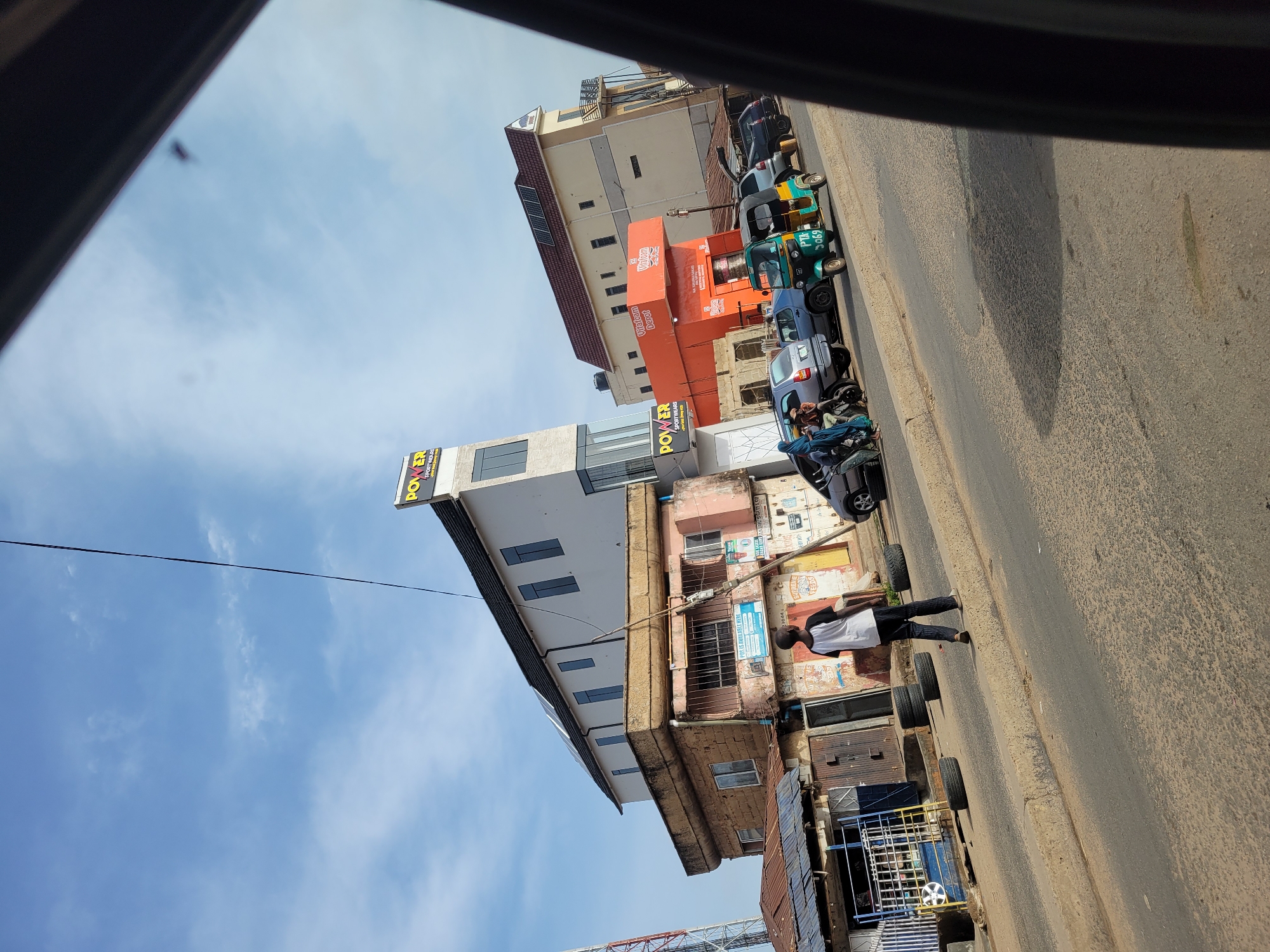 A street scene in Unknown, Unknown is captured from the perspective of someone inside a vehicle. The road runs diagonally from the bottom right to the top left. A man in a white shirt and dark pants walks along the right side of the road, next to stacked tires and metal structures. Buildings line the left side of the road, including structures with signs like "POWER" visible. Several vehicles, including cars and auto rickshaws, are parked along the left side. The sky is visible above the buildings, with some clouds present. A portion of the vehicle interior is visible at the top of the frame.