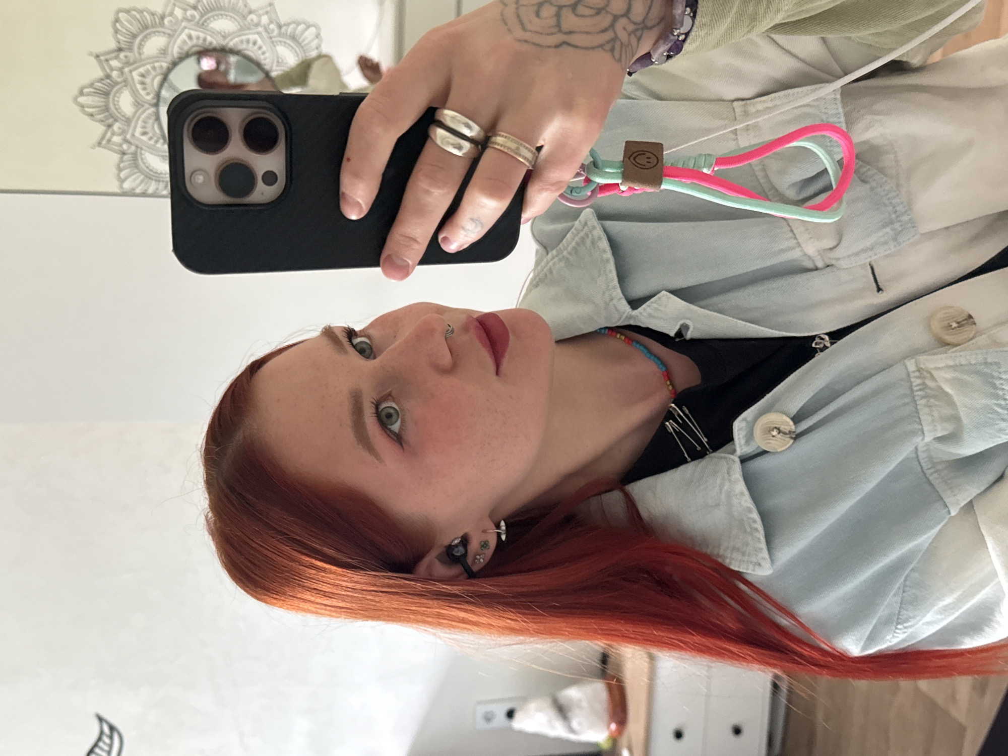 A woman is taking a selfie indoors, presumably in a residential space. She has red hair, fair skin with freckles, and green eyes. She has multiple piercings in her ear and a piercing on her upper lip. She's wearing a light blue denim jacket over a black top, and a beaded necklace. On her fingers, she wears two prominent rings. She's holding a black smartphone with a multi-camera system. A wrist strap is attached to the phone. The background includes a decorative mirror, a white wall, and a glimpse of furniture. The location in the city of Rostov-on-Don, Russia is not directly discernible from the provided image.
