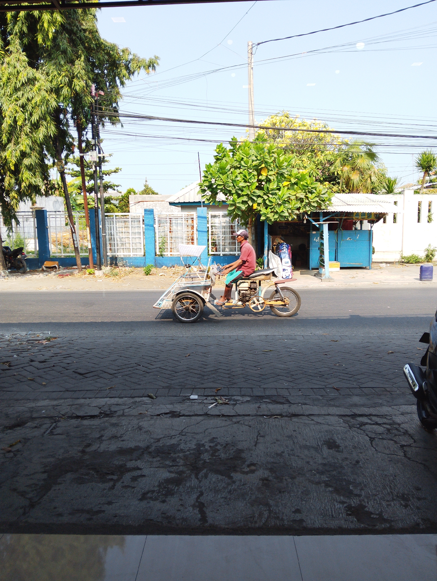 A man wearing a red t-shirt, blue shorts, and a white cap rides a three-wheeled motorized vehicle down a paved street in Manaruwi, Indonesia. The vehicle has a yellow frame, a single rear wheel, and a passenger seat covered with a white cloth. The man is positioned on the driver's seat, which is attached to the vehicle's engine. The street is lined with a blue fence, a sidewalk, and buildings with white walls and blue trim. Trees with green foliage are visible on the left and right sides of the image. The sky is clear and blue, with a few power lines strung across it. The scene appears to be a typical street view in a residential or commercial area.