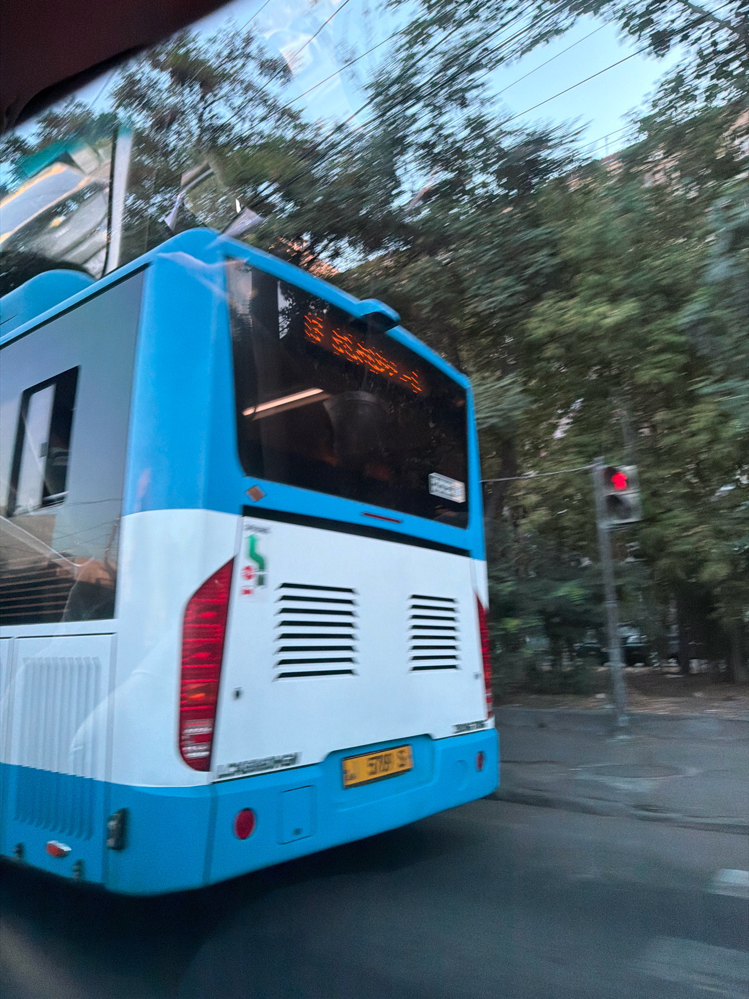 The image shows the rear of a white and blue bus in Yerevan, Armenia. The bus has a blue bumper and a yellow license plate with the inscription "5759 S". The bus has red taillights and a white horizontal vent on the rear. The bus is moving, as indicated by the motion blur of the surrounding trees and the sky. A traffic light with a red signal is visible in the background to the right of the bus. The scene appears to be a street in a residential or urban area, with trees lining the road. There are no visible people in the immediate vicinity of the bus. The overall lighting suggests it is daytime, possibly late afternoon or early evening.