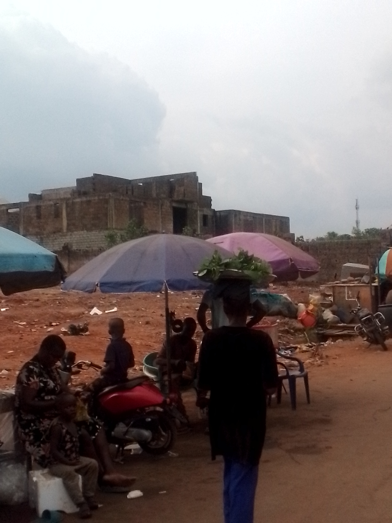 The image shows a street scene in Orlu, Nigeria, on an overcast day. In the foreground, a woman sits on a white block with a child seated in front of her. Behind them, a red scooter is parked. Several people are gathered under a blue umbrella. A person with a load of green plants on their head walks away from the group toward the viewer. Another purple umbrella is visible further in the background, where trash accumulates. A building under construction stands in the distance to the left. A cell phone tower can be seen behind a tree line on the right. The ground is reddish-brown.