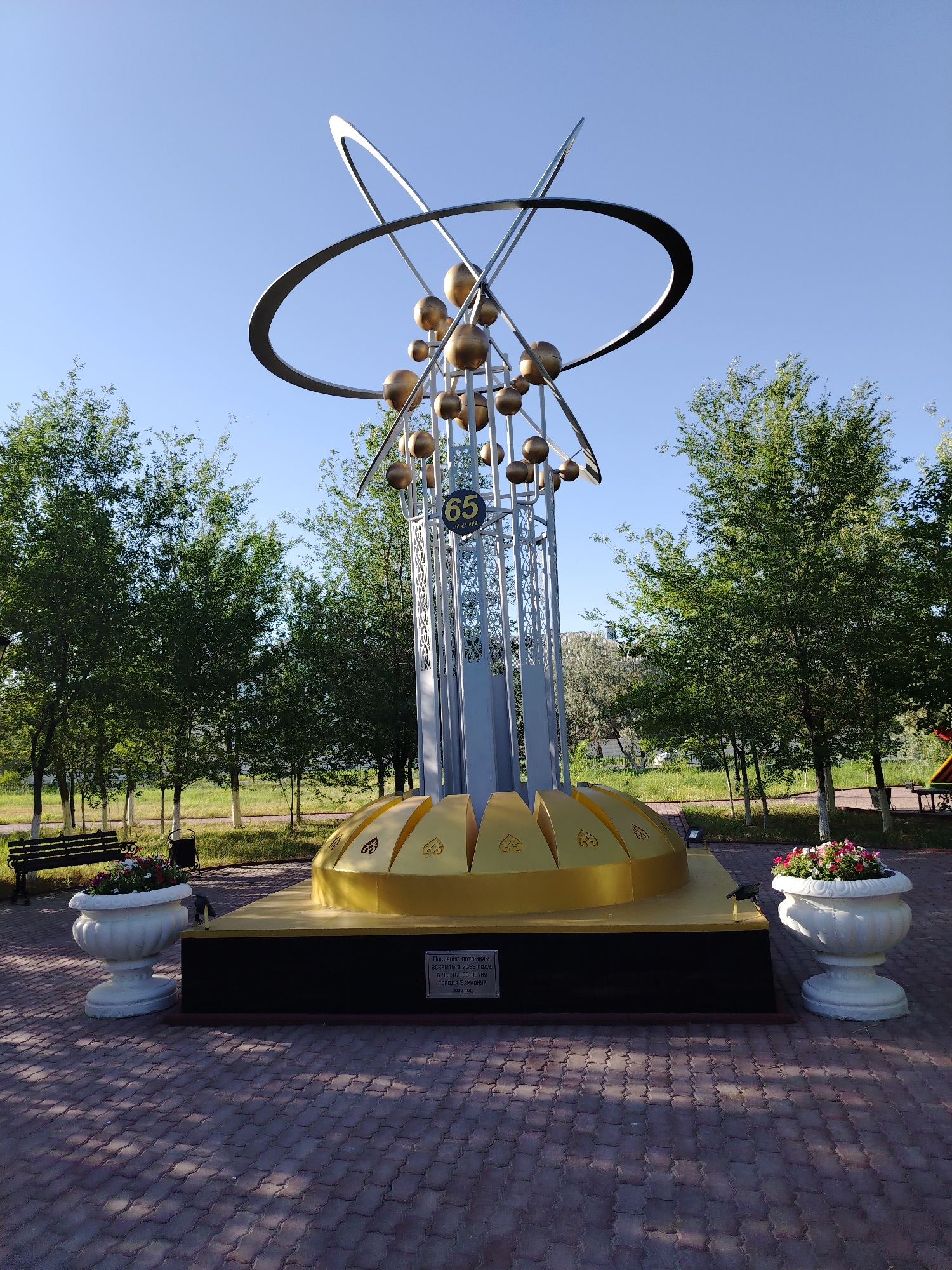 The image shows a monument with a globe-like structure at the top, supported by a silver-colored lattice tower, set on a golden base. The monument features metallic spheres and rings at the apex. A circular emblem with the number "65" is visible on the tower. The monument is located outdoors on a paved area, surrounded by trees and a clear blue sky. There are two large white planters filled with flowers, one on each side of the monument's base. A park bench and a trash can are situated to the left of the monument. The base of the monument has a plaque with text, and the overall scene suggests a public park or memorial area in the city of Baikonur, Kazakhstan.