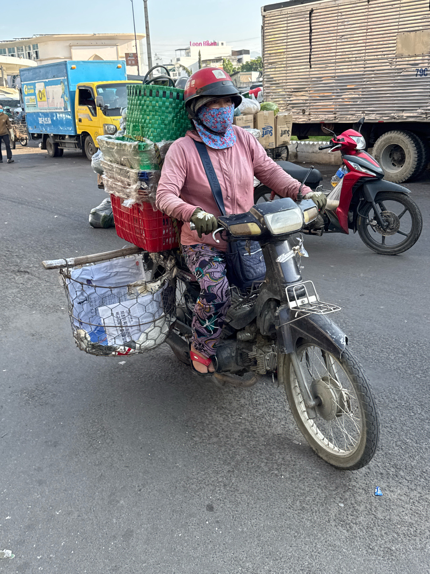 An overhead shot shows a person riding a dark-colored motorcycle in what appears to be a street in Nha Trang, Vietnam. The rider, wearing a pink long-sleeved shirt, a red helmet, and a blue and white patterned mask, has a large quantity of goods secured to the motorcycle. A red basket is positioned behind the rider, filled with green plastic baskets and various other items. A wire basket hangs off the side of the motorcycle in front of the rider, containing white objects and some paper. The motorcycle is on an asphalt road, with a large silver truck parked on the right side of the image and a yellow and blue truck on the left. The environment suggests a bustling urban setting with commercial activity.