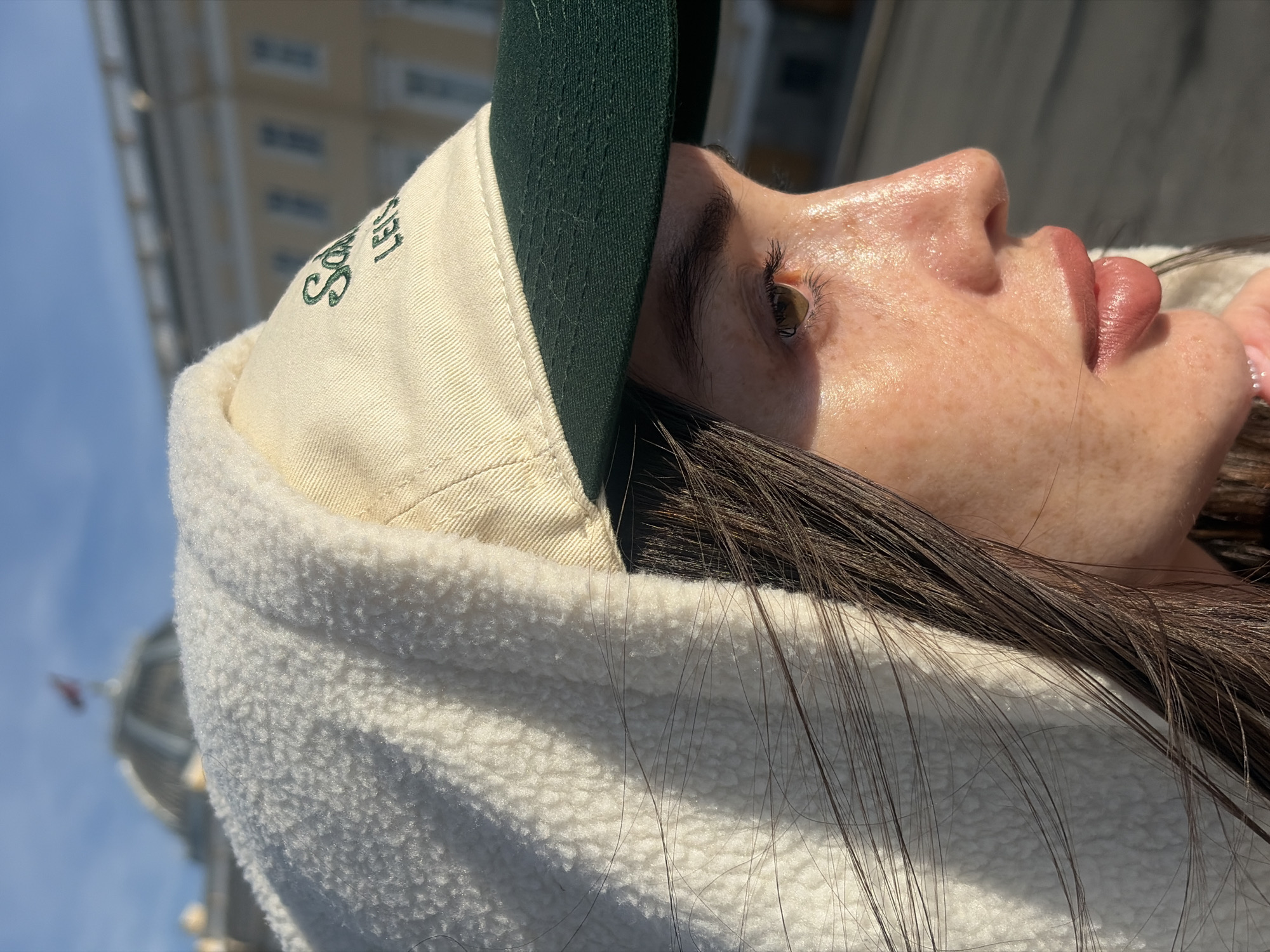 A close-up shot features a young woman's face in profile, her head tilted upwards. She wears a cream-colored baseball cap with dark green trim and embroidered green text. A fuzzy white hood is pulled up around her neck and head, partially obscuring her. Her dark brown hair is visible, falling loosely around her face.
The woman's skin is slightly shiny, catching the sunlight. She has light-colored eyes, dark eyebrows, and a light dusting of freckles across her nose and cheeks. Her lips are slightly parted, revealing a rosy hue. The lighting suggests it is daytime, with bright sunlight casting shadows and highlighting her features.
In the background, a blurred building with multiple windows is visible against a clear blue sky, along with what appears to be the dome of a building, possibly a landmark, peeking out from behind the woman's hood. The scene appears to be outdoors in an urban environment. The crops reveal embroidered text on the cap that reads "SOUL LESS."