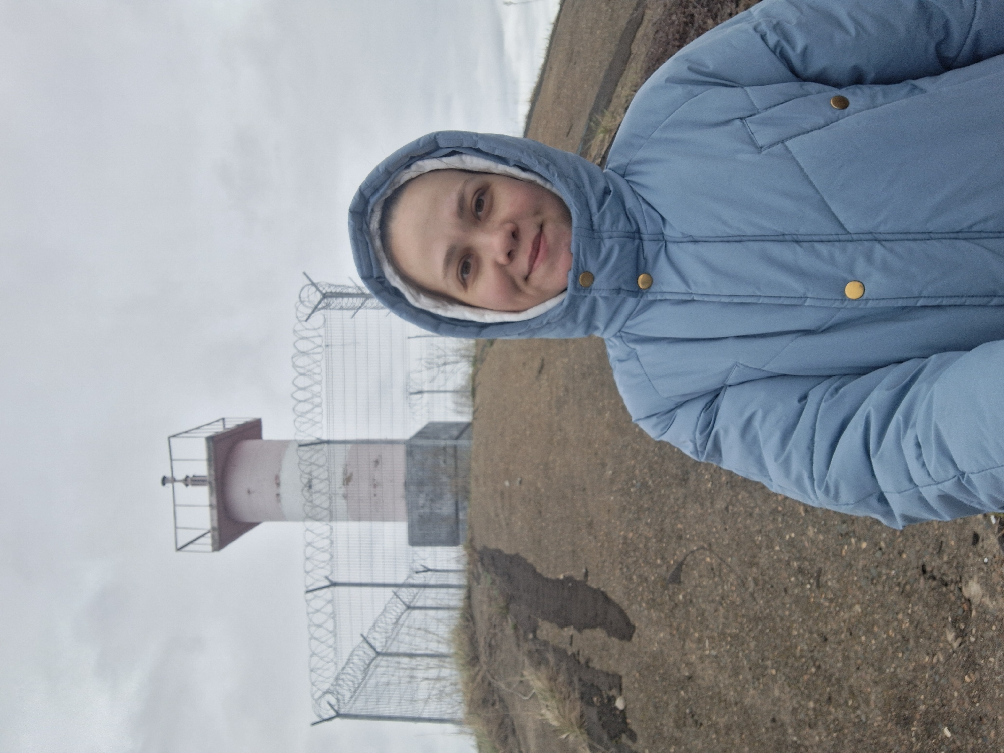 A woman is captured in a close-up outdoor portrait, posing under a cloudy sky in what appears to be Chaikovsky, Russia. She is wearing a light blue, quilted puffer jacket with the hood up, framing her face. Her expression is calm and pleasant, with a slight smile on her lips, and she looks directly at the camera. The jacket features gold-colored snap buttons down the front and on a small chest pocket. The interior lining of her hood is a contrasting white or light cream color.
In the background, under a uniformly grey, overcast sky, a distinctive structure resembling a small lighthouse or a navigational beacon stands. This structure has a light-colored, possibly pale pink or white, cylindrical base and a darker, rectangular top section with a metal framework and an apparatus at its peak. In front of this structure and extending across the mid-ground is a chain-link fence topped with coils of barbed wire, suggesting an area of restricted access or a boundary. The foreground and mid-ground consist of rough, earthy ground, possibly a dirt path or gravelly terrain. The overall ambiance suggests a cool, perhaps damp, and overcast day. There is no visible text in the image.
