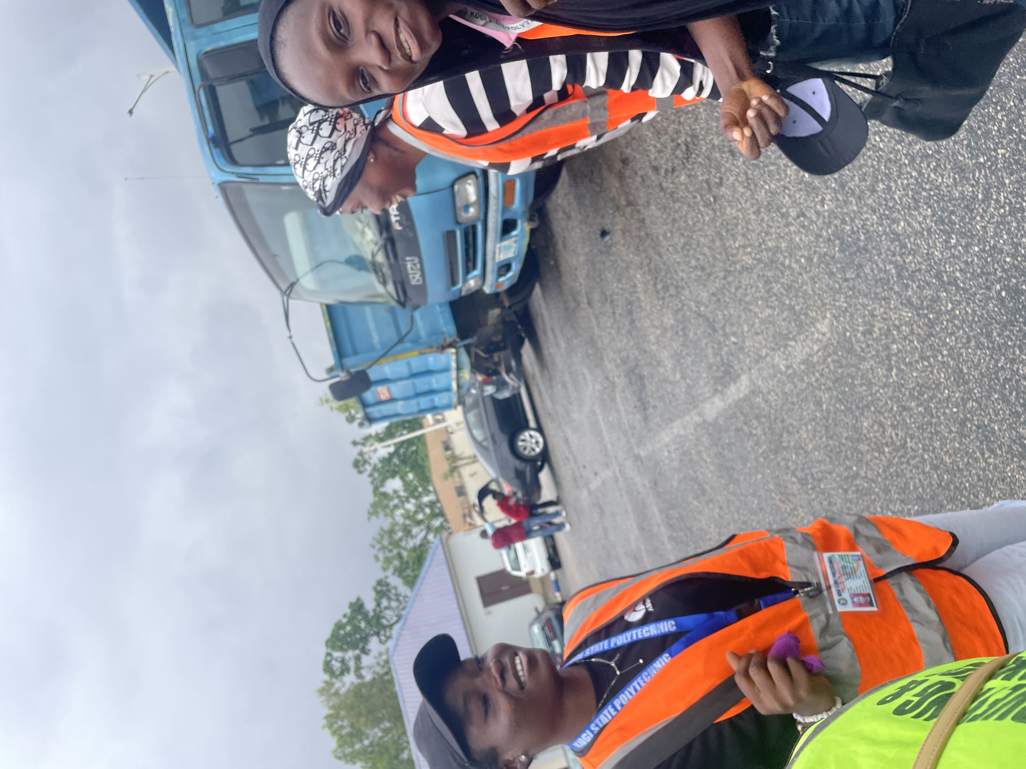 The image depicts an outdoor scene, likely in Lokoja, Nigeria, under an overcast sky. In the foreground, three women are visible. The woman on the left, wearing a black cap and an orange reflective vest, is looking to her left and smiling. A lanyard with a badge is attached to her vest. To her right, another woman, wearing a black and white striped shirt and an orange reflective vest, is also looking to her left and smiling. She has a patterned headscarf on. Partially visible on the far right is a third woman, wearing a black hijab and a black garment. She is looking towards the camera and smiling.
Behind the women, a bright blue Isuzu truck with an open bed is parked. In the background, several other vehicles are present, including a dark-colored sedan and a white car. There are buildings with pitched roofs, some with white facades, and trees with green foliage. The ground is asphalt. The overall impression is a casual gathering of individuals in an outdoor setting, possibly related to work or an event involving the truck.