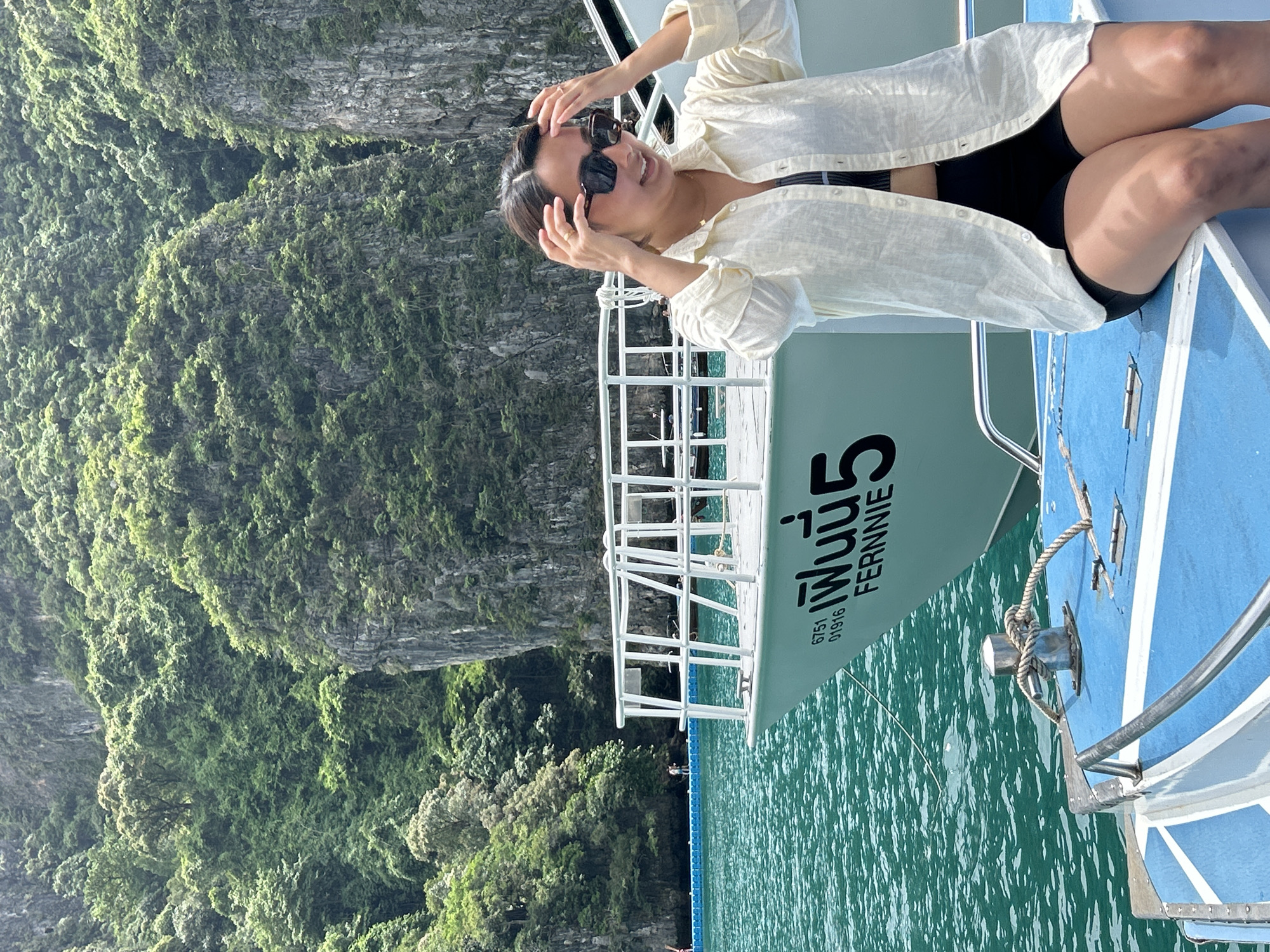 A woman is sitting on the front of a boat in Ao Nang, Thailand, with a boat behind her. The woman is wearing a light-colored, unbuttoned shirt over a black bikini top and black shorts. She has dark hair and is wearing sunglasses. She is looking to her right, with her hands on her head. The boat behind her is white with the name "FERNNIE" written on the side in black lettering, along with some Thai text and numbers. The water is turquoise and calm. In the background, there are steep, rocky cliffs covered in lush green vegetation. A blue floating walkway is visible to the left of the boat, with a few people walking on it. The sky is overcast, but the light is bright.