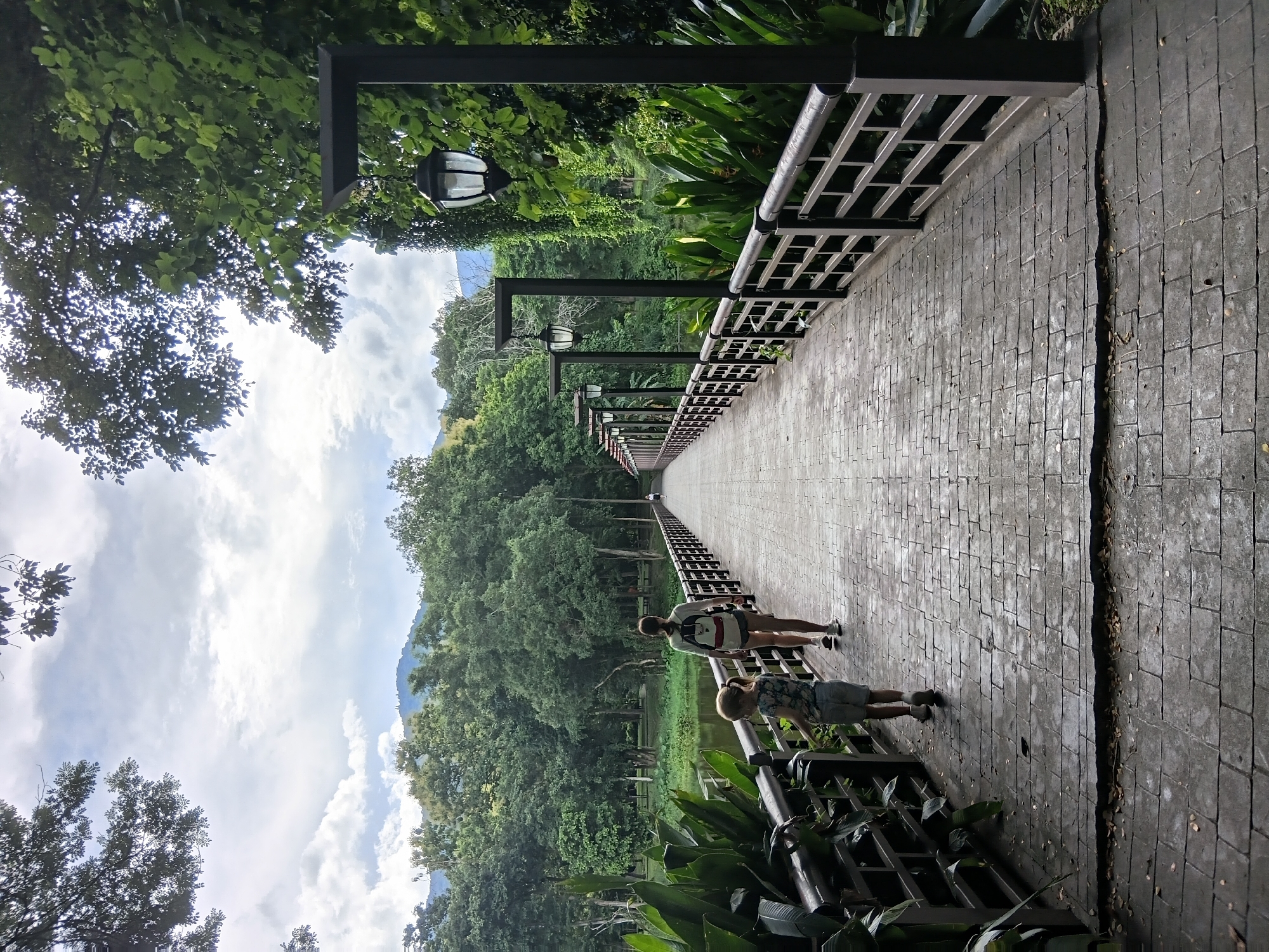 A woman and a child walk away from the camera on a paved bridge lined with lampposts and railings. The woman, wearing a backpack, and the child, in a floral shirt and shorts, are walking towards the center of the image. Lush green foliage and trees surround the bridge, with a body of water visible to the left. In the background, a mountain range is visible beneath a cloudy sky. The scene appears to be a park or a scenic walkway within the city of Chiang Mai, Thailand.