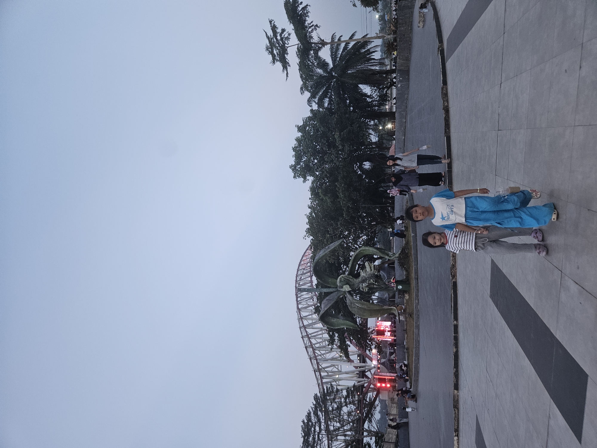 The image depicts a wide outdoor paved area in the city of Teluk Dalam, Indonesia. In the foreground, two young girls are walking towards the viewer. The girl on the left wears a striped t-shirt and grey pants, while the girl on the right wears a white and blue t-shirt with blue pants. Behind them, other individuals are visible walking in the background.
Dominating the middle ground is a large, abstract metal sculpture, possibly depicting a stylized flower or plant. Behind the sculpture, a red and white truss bridge spans across the scene. A stage with bright red lights and tents is set up below the bridge, suggesting an event or performance is taking place. Numerous trees, including palm trees, are scattered throughout the landscape, and a general hazy or overcast sky covers the upper portion of the image. The overall lighting suggests it is either early morning or late evening.